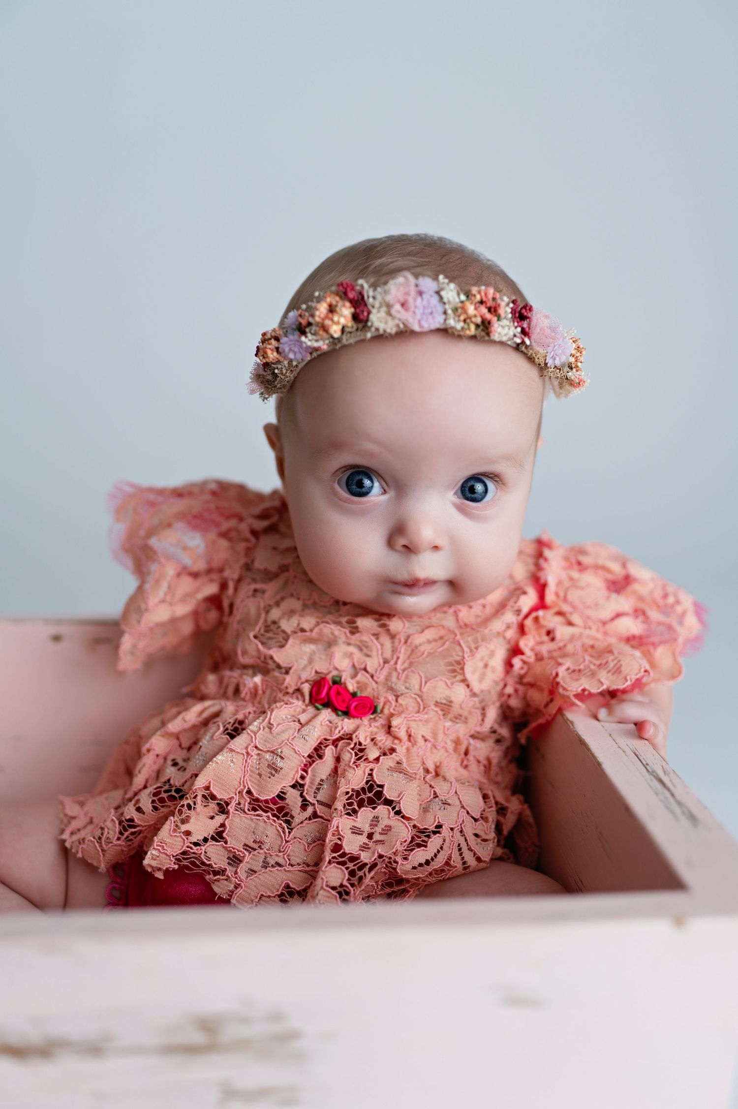 A baby wearing a peach lace dress and floral crown poses for a professional photography session.