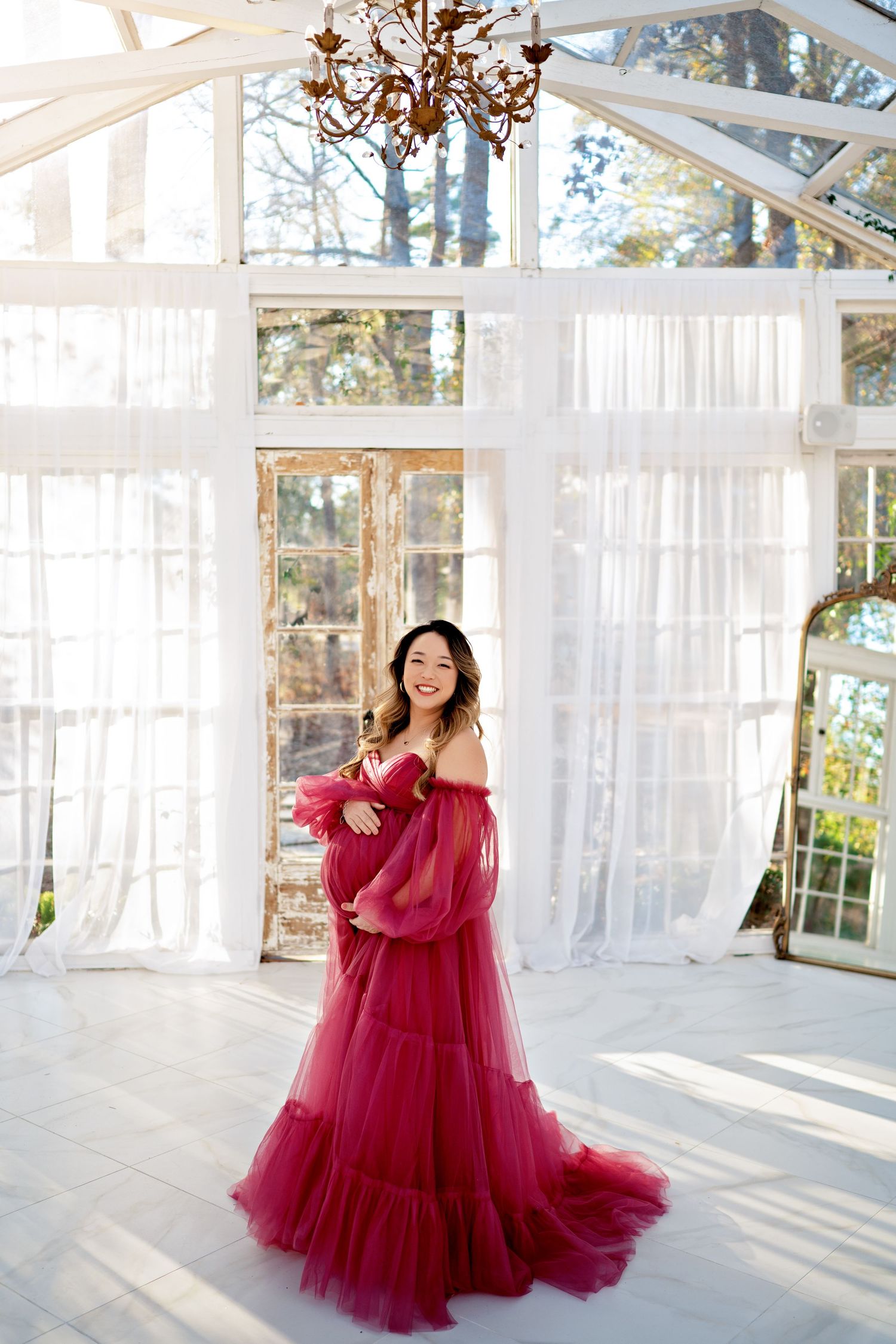 Ethereal burgundy tulle gown flows gracefully in a bright sunlit room with white sheer curtains and wooden beams.