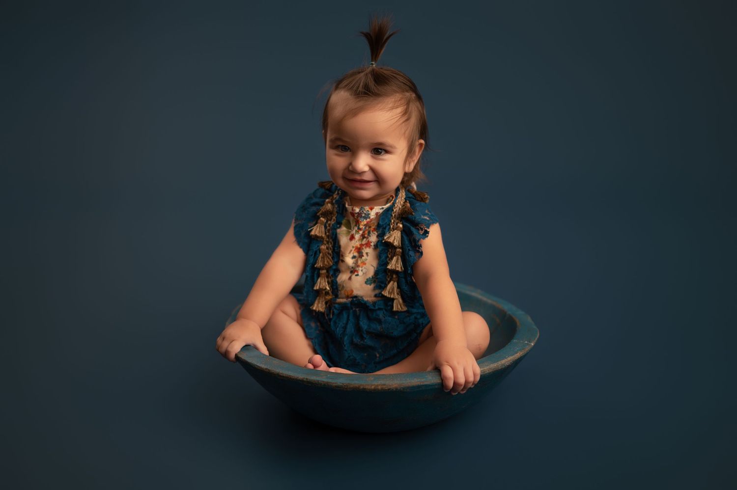 A young child in a navy blue dress sits in a round metal basin against a dark teal backdrop.