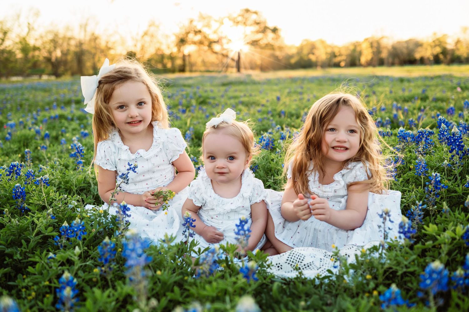 Children in white dresses sitting together among vibrant blue wildflowers during a golden sunset photo session.
