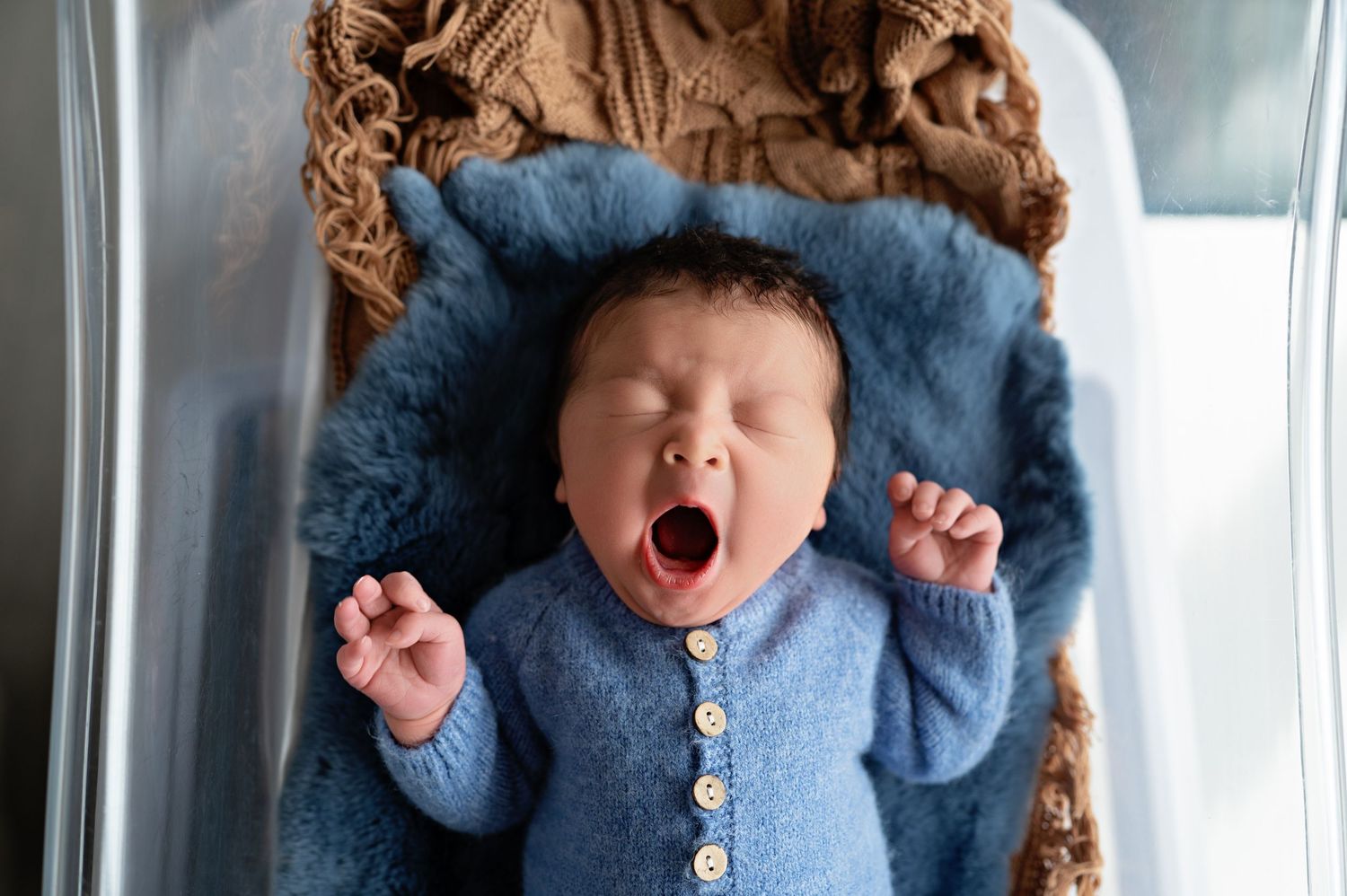 Baby in blue knit cardigan yawning while lying on soft blue blanket with brown plush toy.