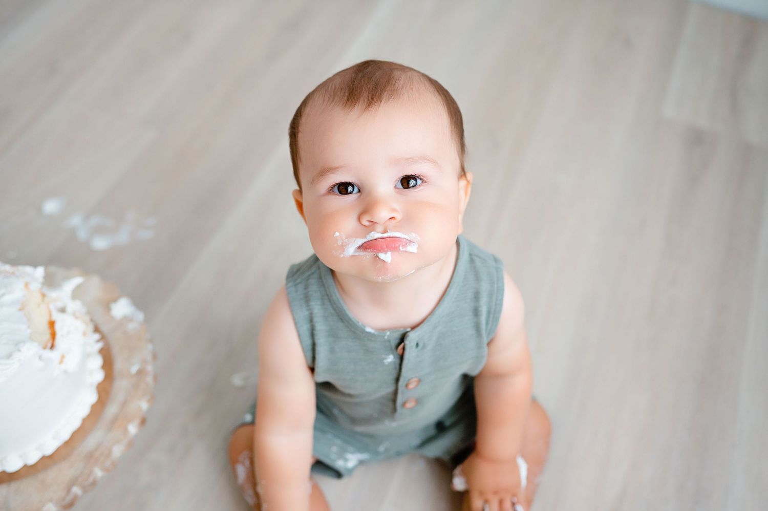 Baby in teal outfit with messy face sitting on floor near cake.