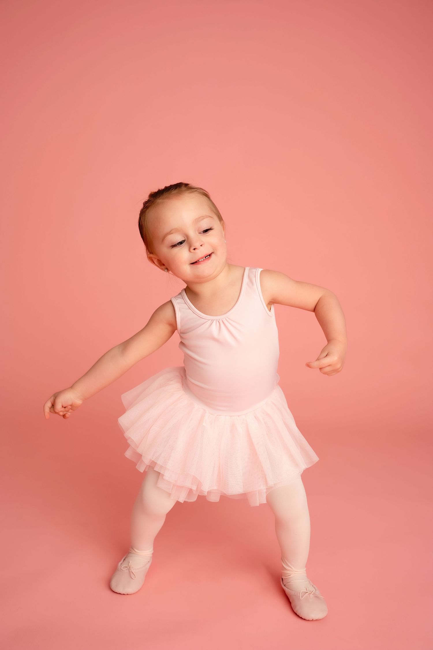 Small ballerina practicing dance moves in pink tutu against coral wall.