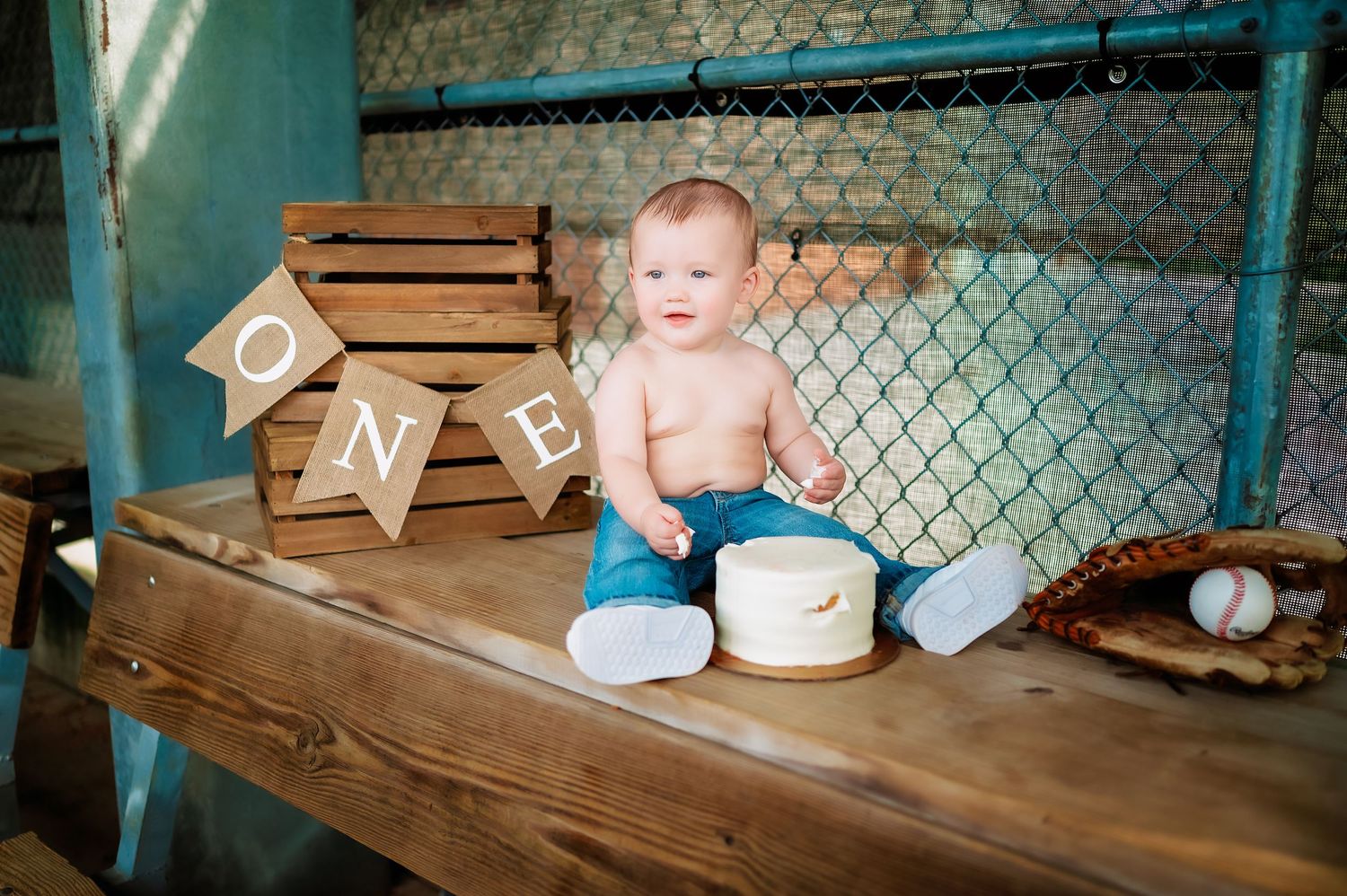 A baby sits on a wooden bench with baseball decorations and ONE letter blocks.