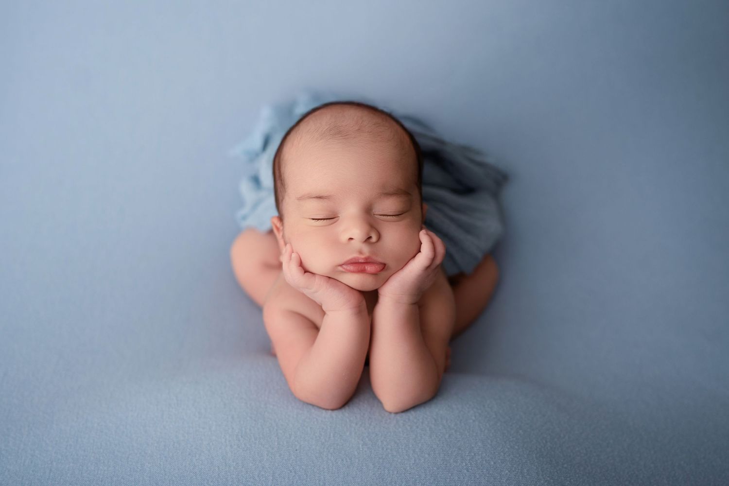 Peaceful newborn baby sleeping with hands under chin on soft blue blanket in professional photography pose.