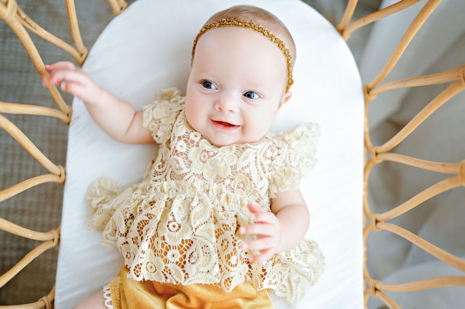 Baby in delicate ivory lace dress lying on white blanket with natural light streaming through branches.