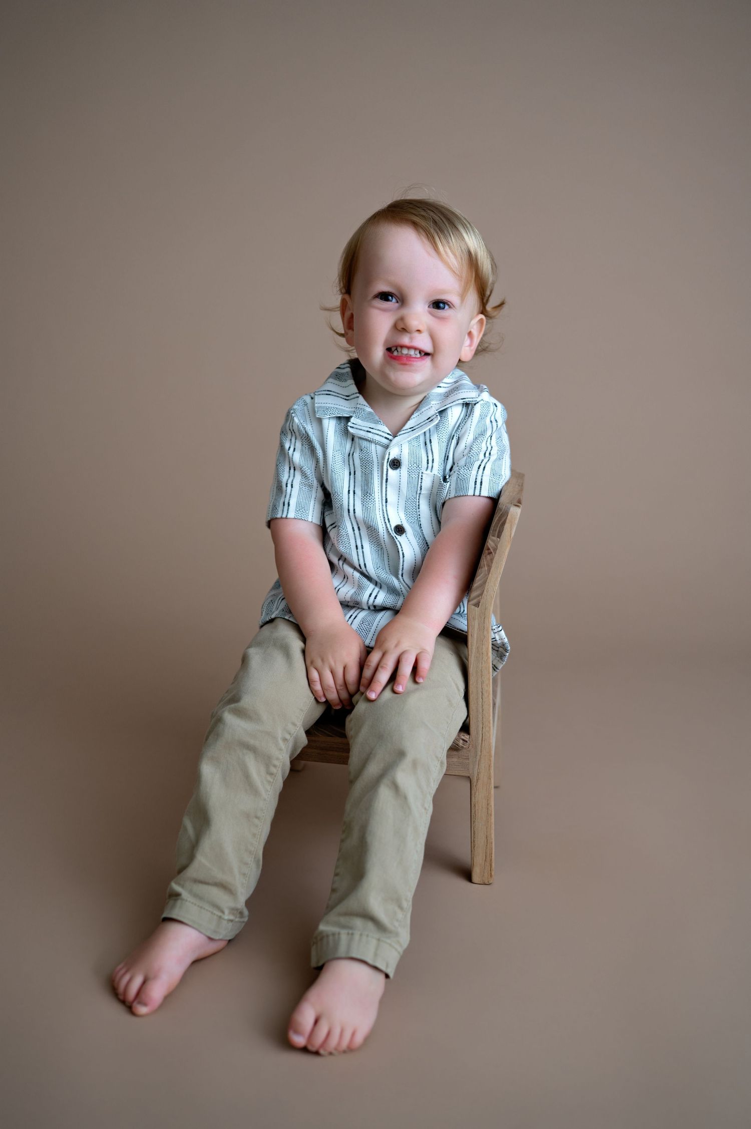 Small child sits in wooden chair wearing striped button-down shirt and khaki pants against neutral beige backdrop.