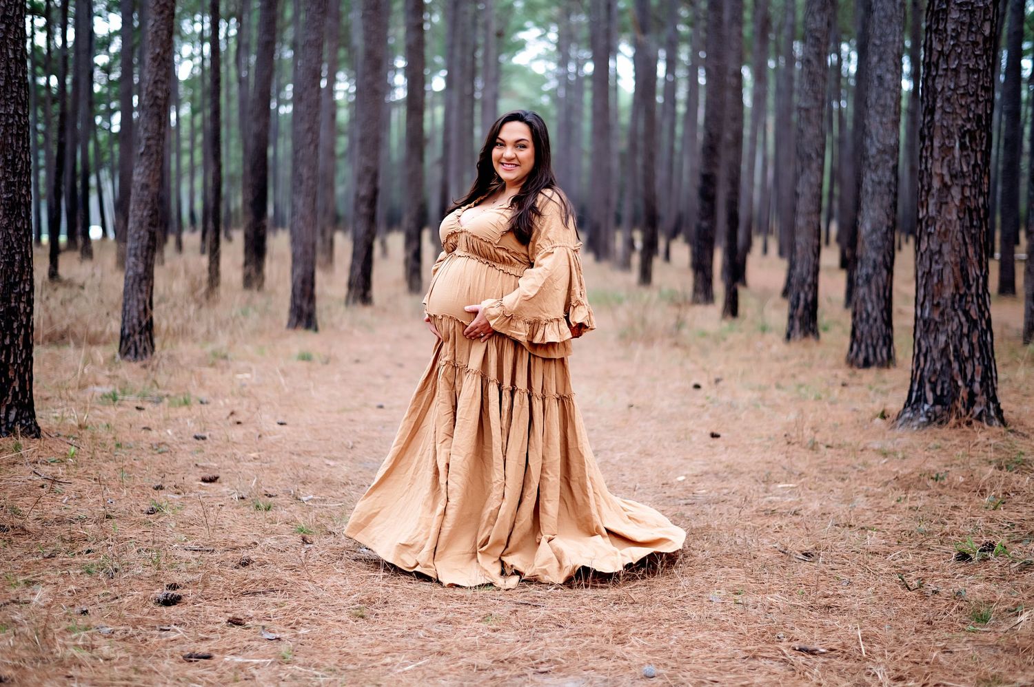 Dramatic maternity portrait in champagne gown among tall pine trees.