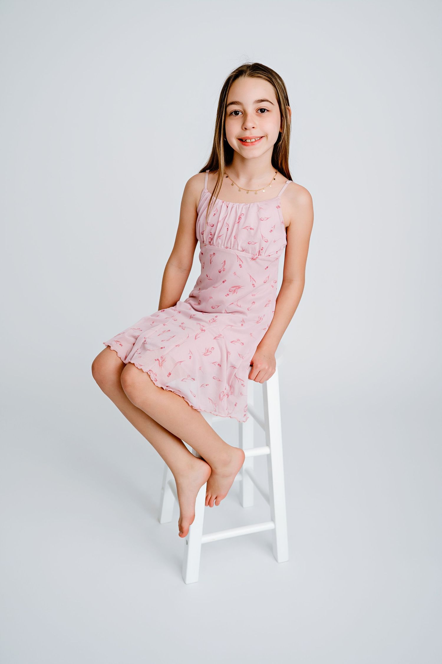 A girl in a pink lace dress sits barefoot on a white stool against a light background.