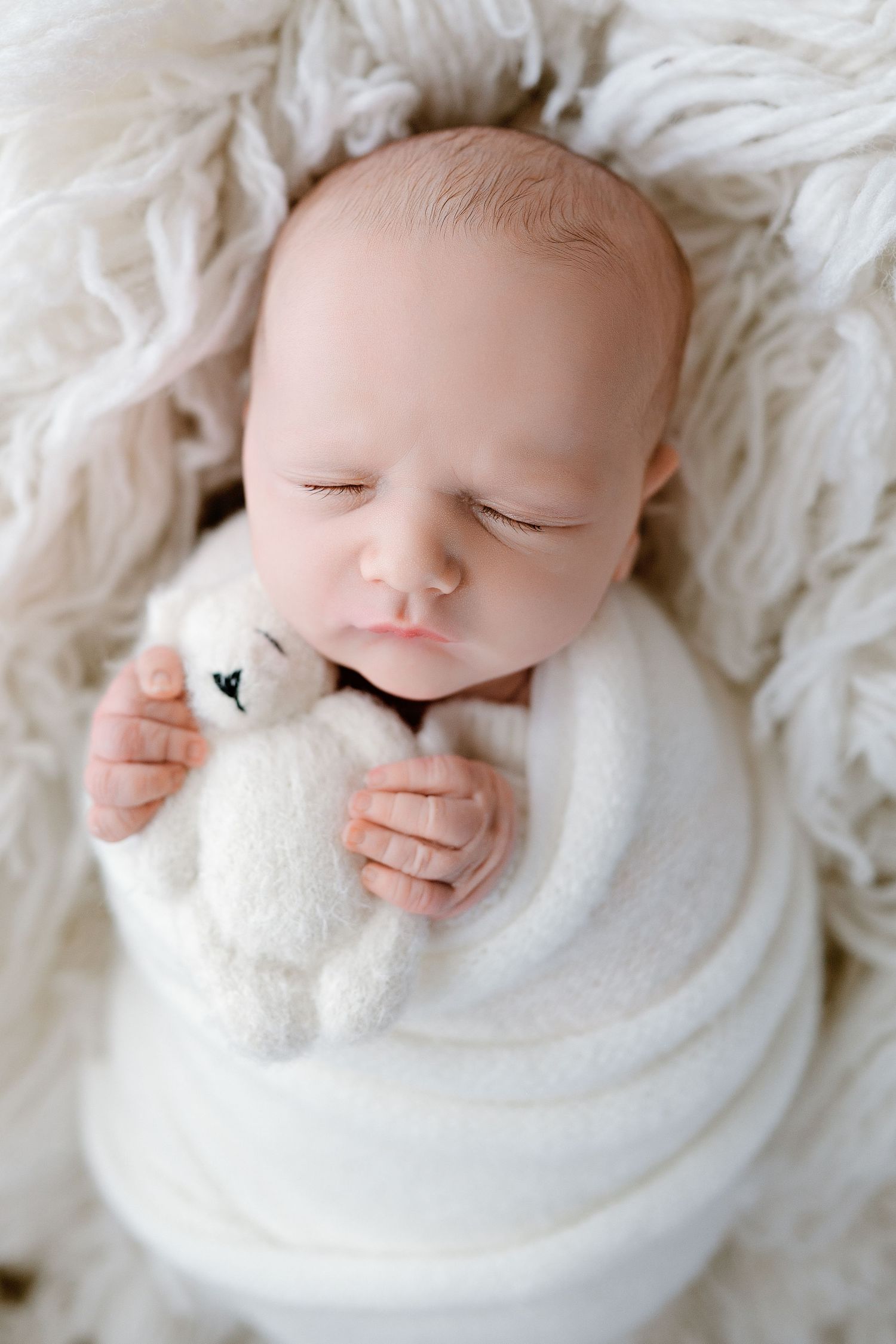 Sleeping newborn wrapped in white swaddle holding small plush toy on fluffy white blanket.