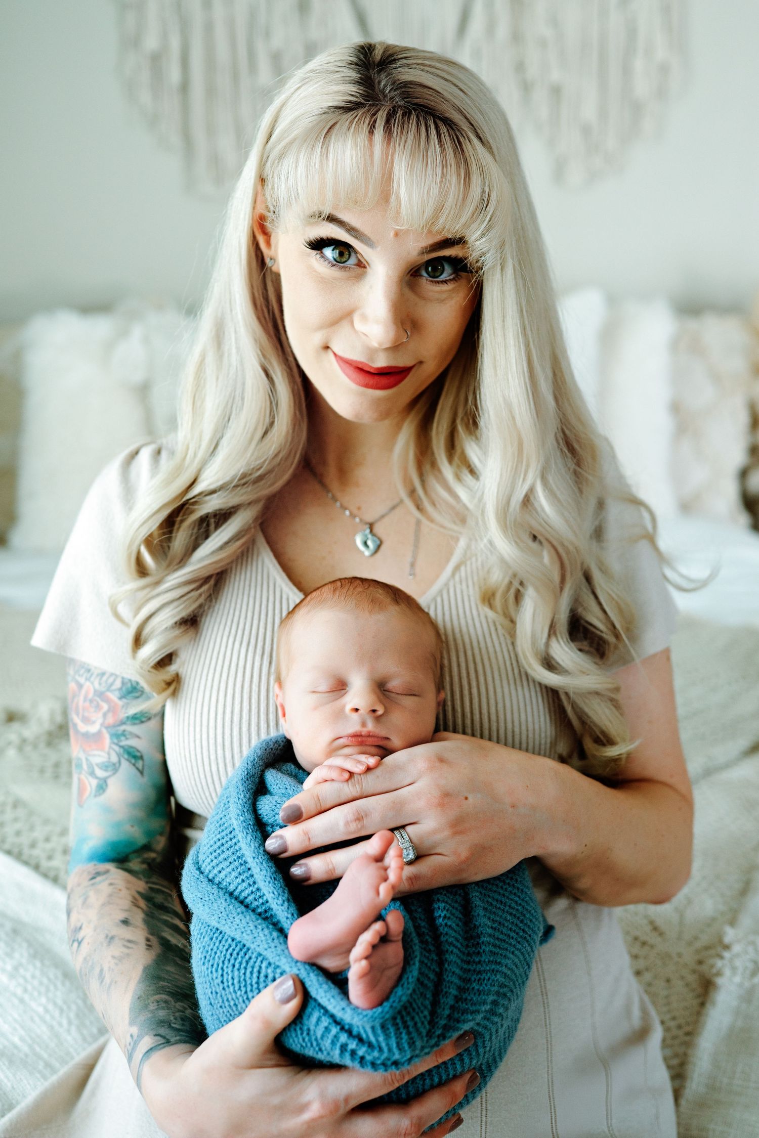 Mother with platinum blonde hair and tattoos holds sleeping newborn baby wrapped in blue blanket while sitting indoors.