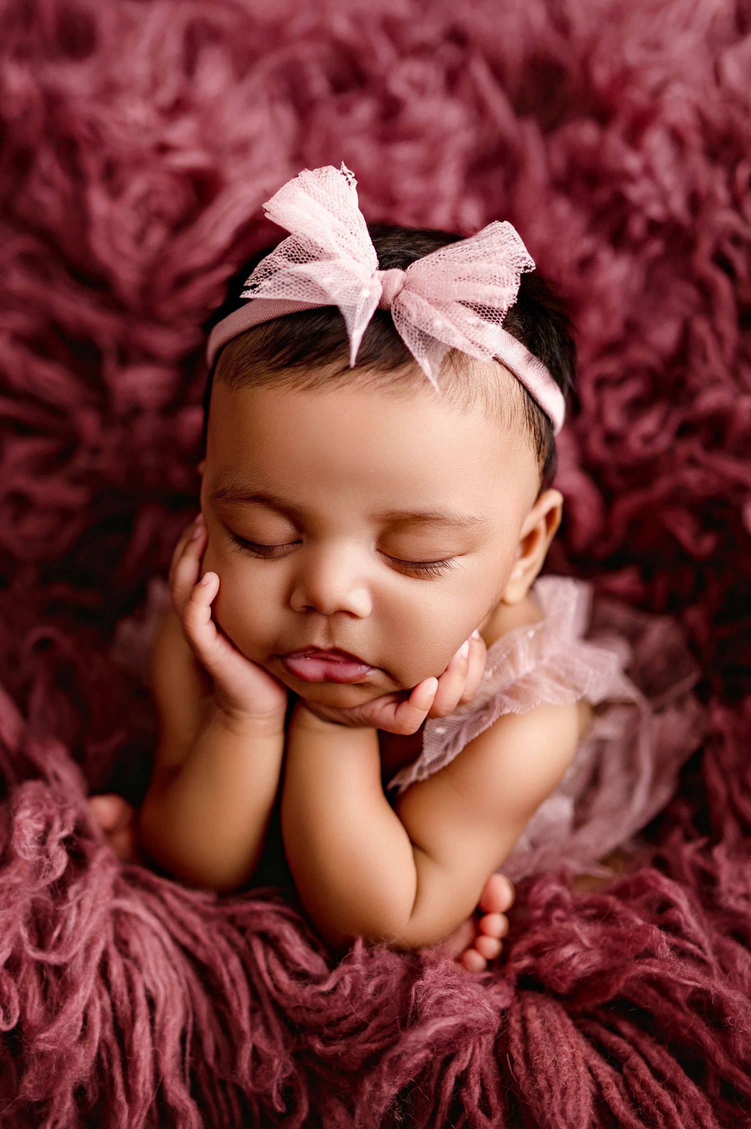 Sleeping newborn baby girl in pink tutu and bow headband posed on mauve flur rug in professional photo.