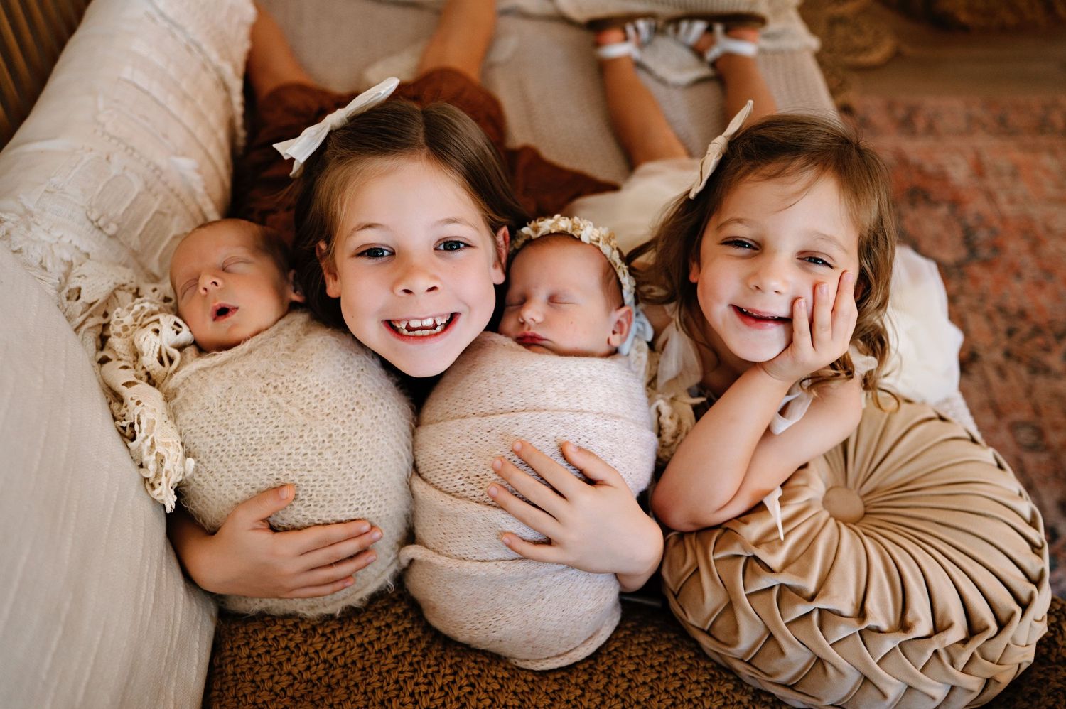 Two smiling girls pose with twin newborns wrapped in cream swaddles on a cozy bed.