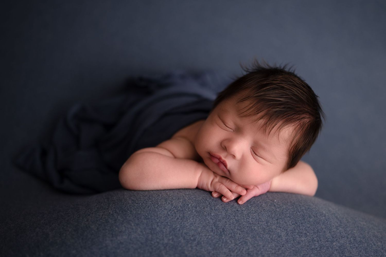 Peaceful sleeping newborn baby in dark grey setting rests with hands folded under head.