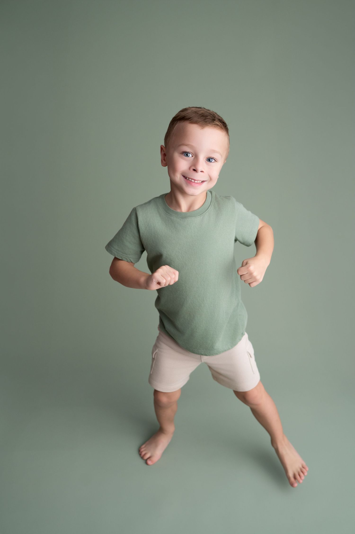 Energetic child in sage t-shirt and shorts jumps and poses against soft green studio backdrop.