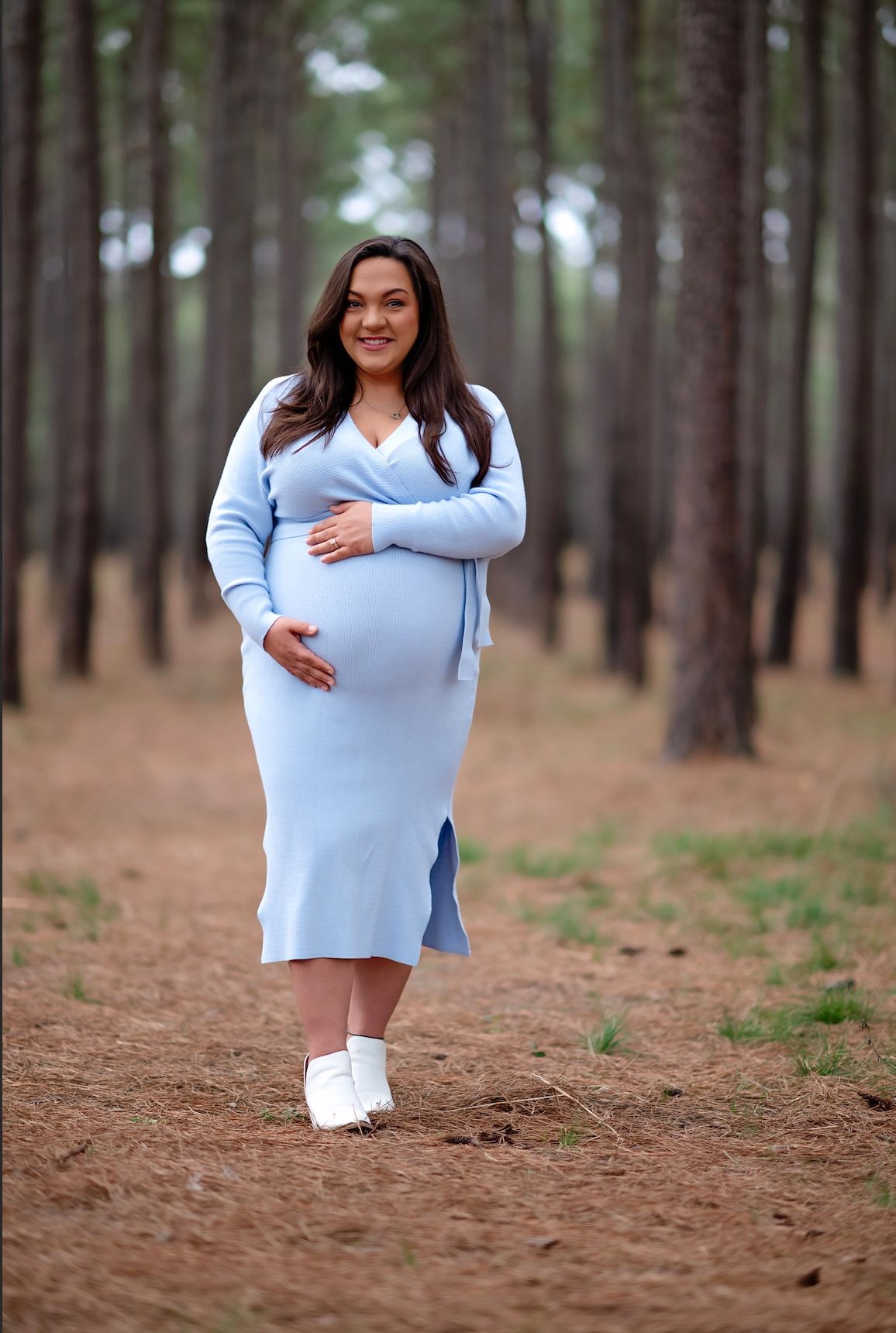 A maternity portrait in a blue knit dress on a path through a pine forest.
