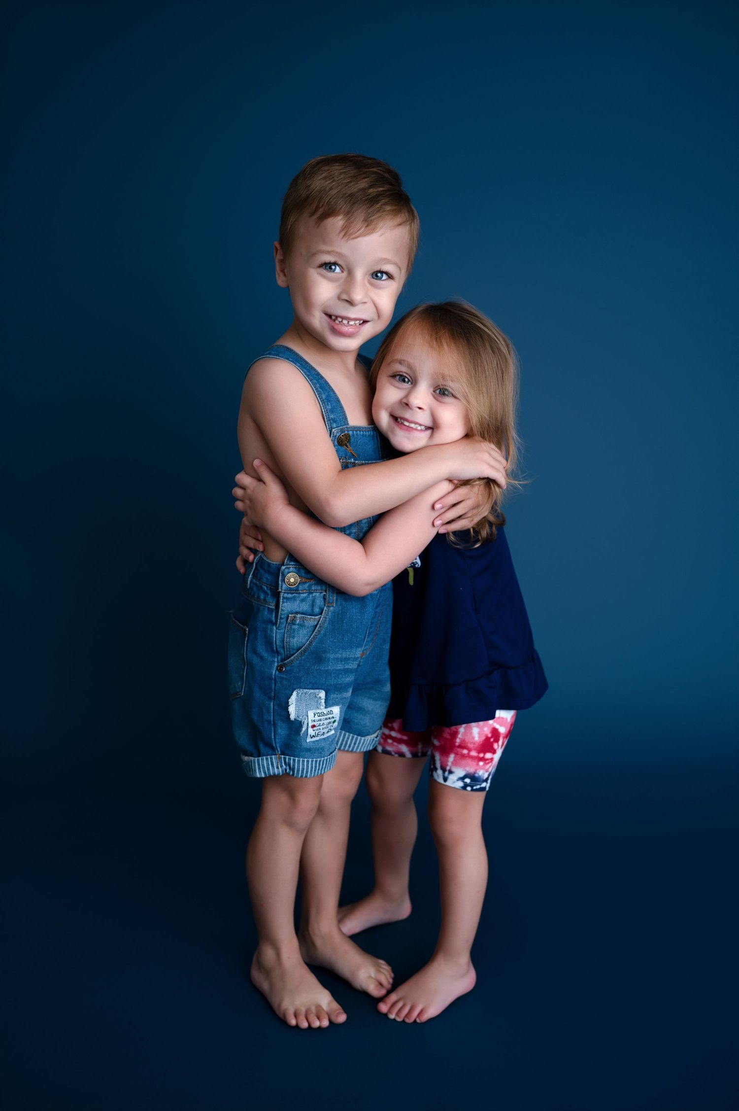 Two young children in summer outfits hugging and smiling against a dark blue background.