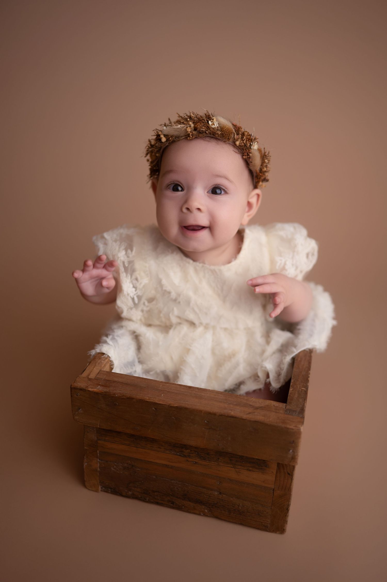 A baby wearing a cream lace dress smiles while lying in a rustic wooden crate against a brown backdrop.