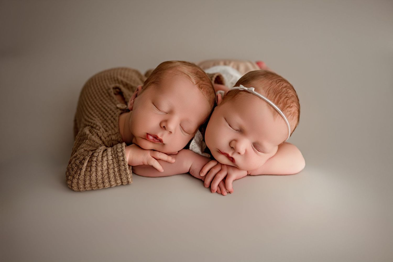 Two sleeping newborn twins posed side by side, wearing knit outfits, resting chins on hands against a neutral background.