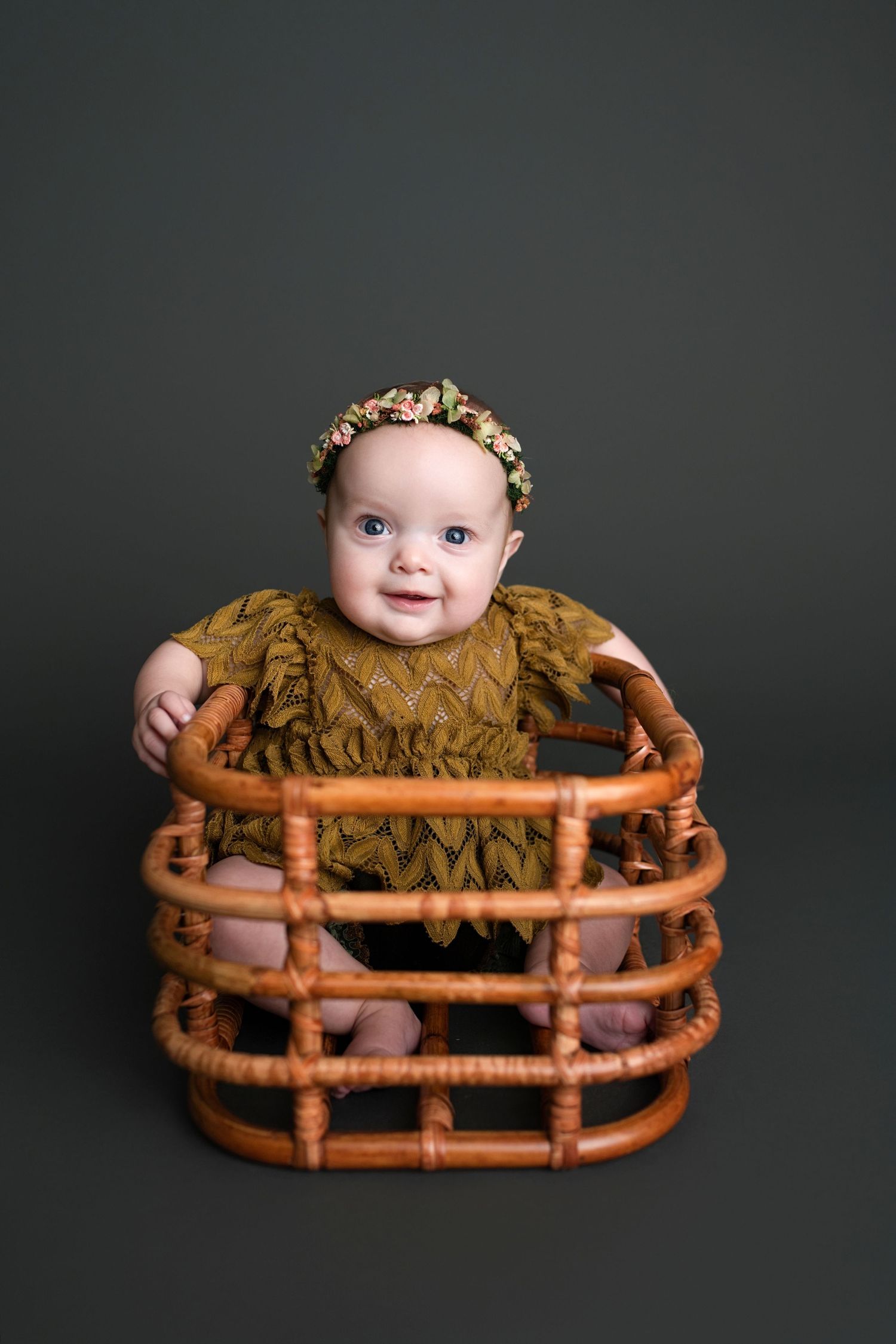 A baby wearing a floral crown and olive green dress sits in a vintage rattan basket chair against a dark background.