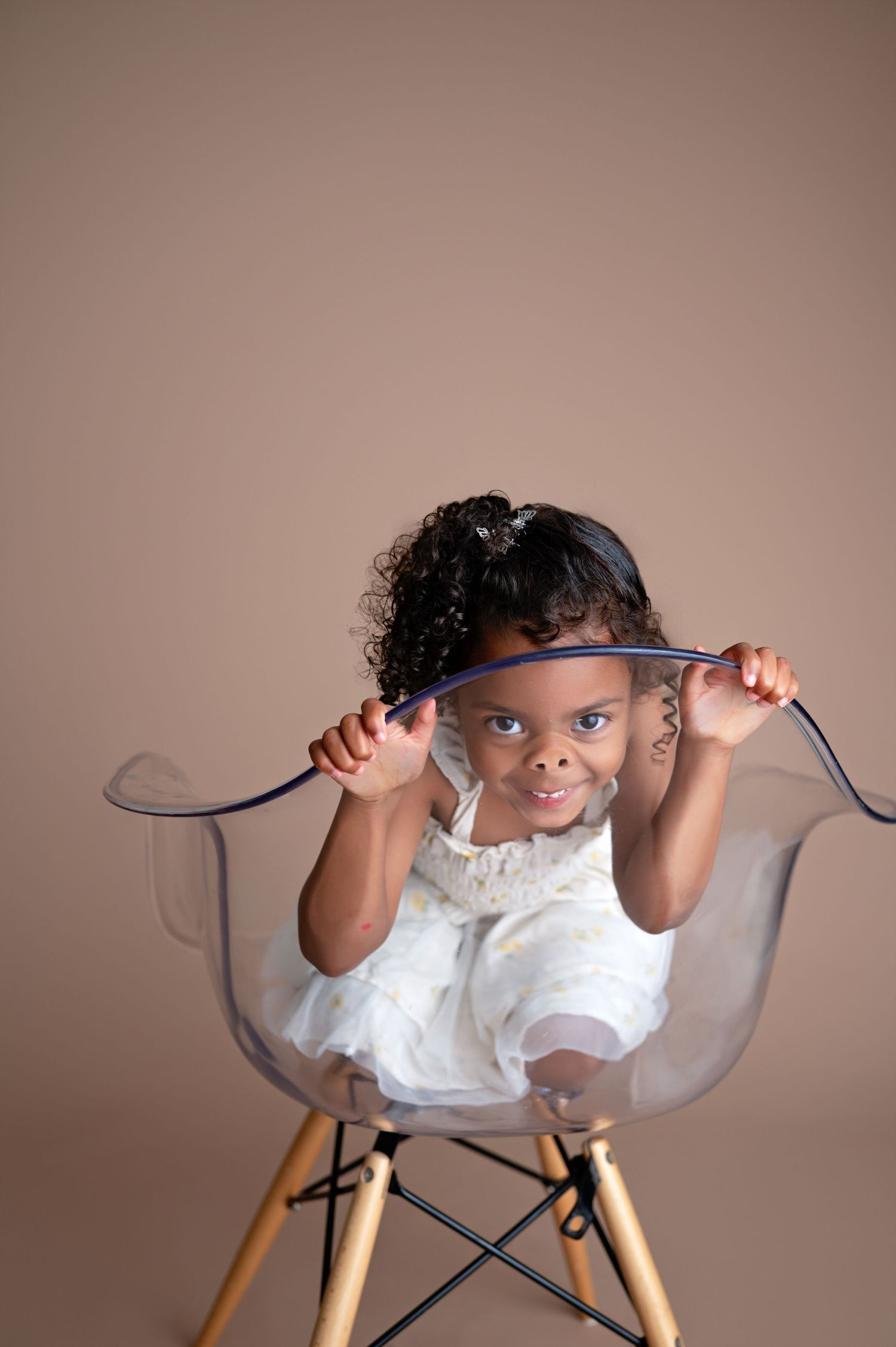 A young child in a white dress sits playfully in a modern clear acrylic chair with wooden legs.