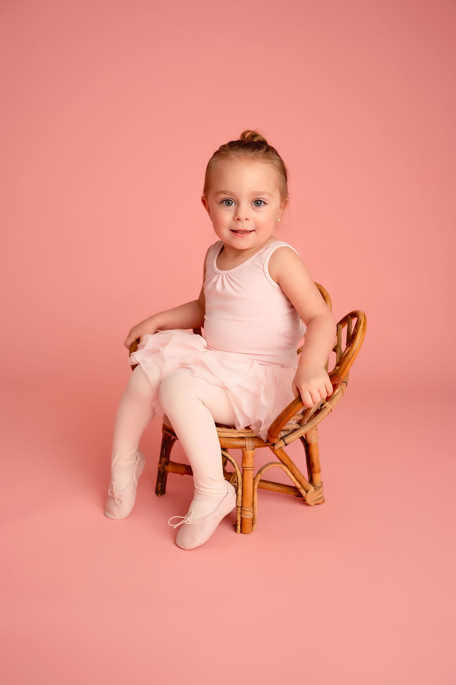 Baby in pink outfit sitting on small wooden chair against coral background.