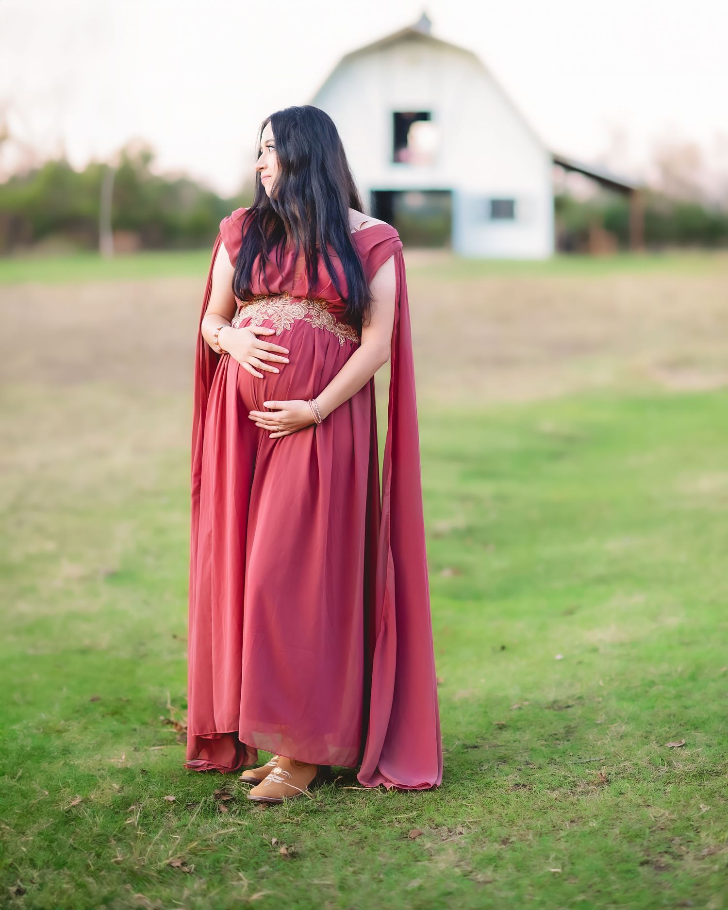 A side view maternity portrait with a red dress and cape in front of a white barn.