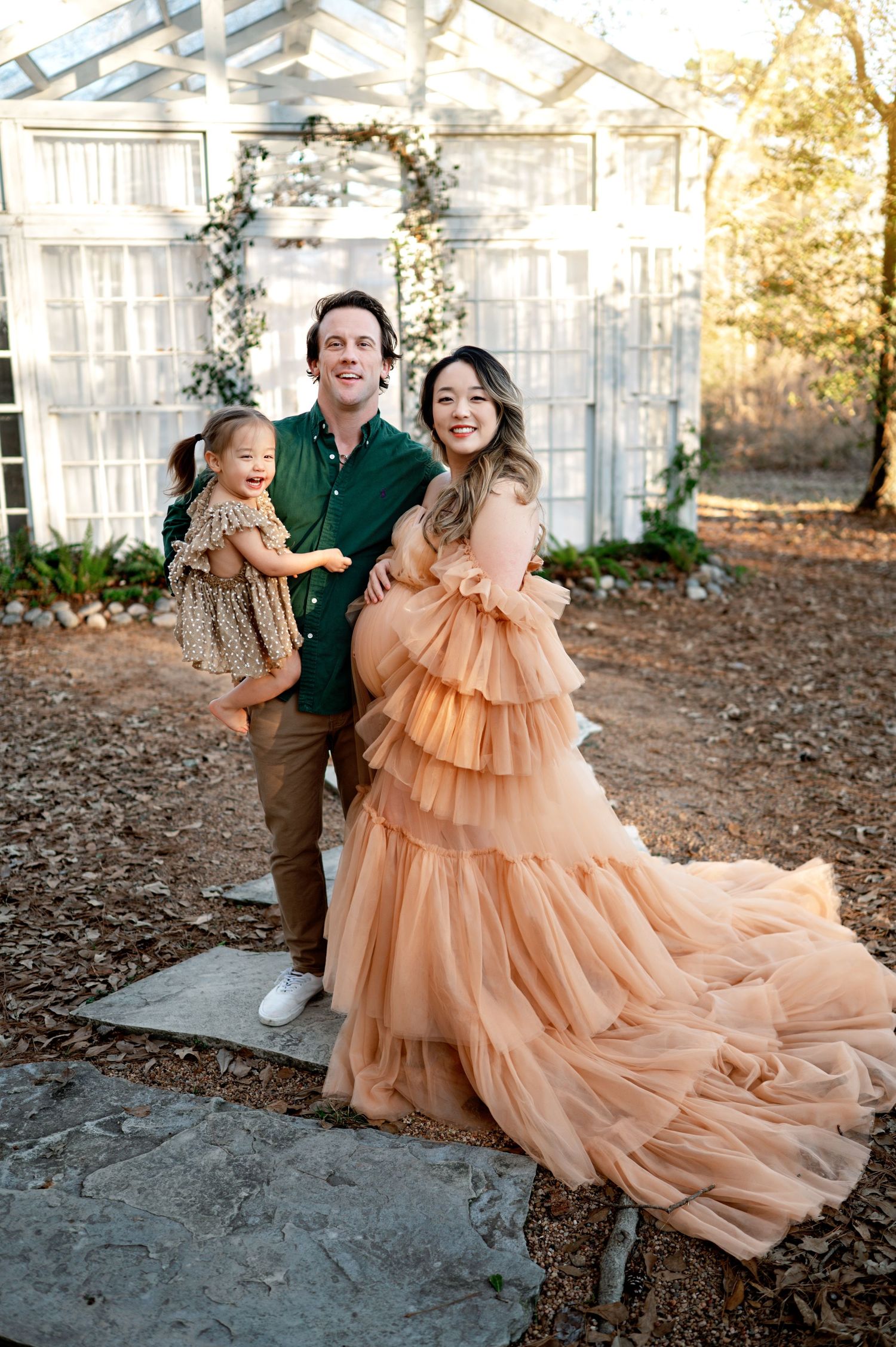 A family poses together in front of a greenhouse, with the woman wearing a flowing peach-colored tiered tulle gown.
