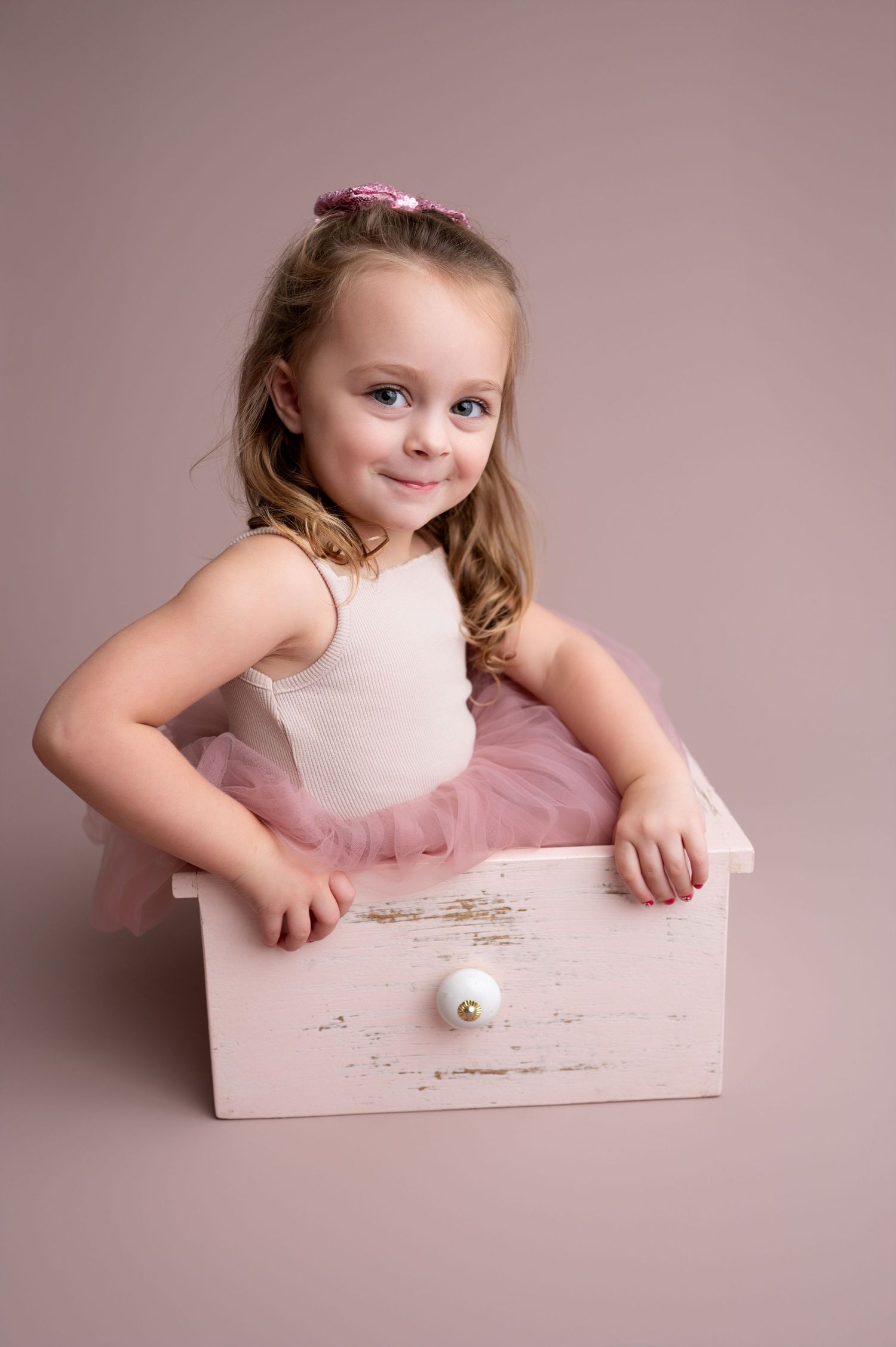 Little ballerina in pink tutu leans playfully on white wooden prop during dance photo session.
