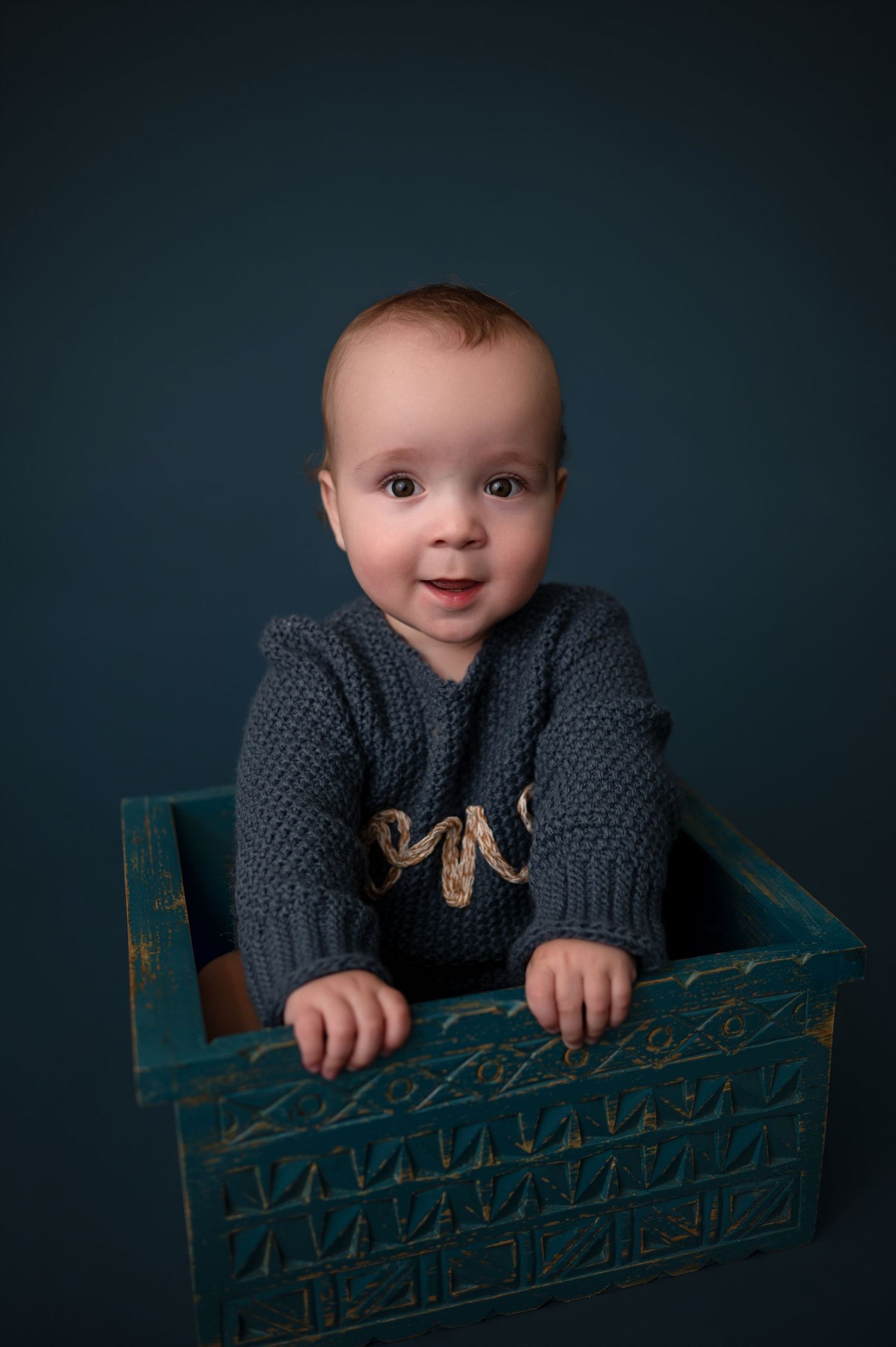 Baby wearing a knit gray sweater peeks over a turquoise vintage crate against dark background in studio portrait series.
