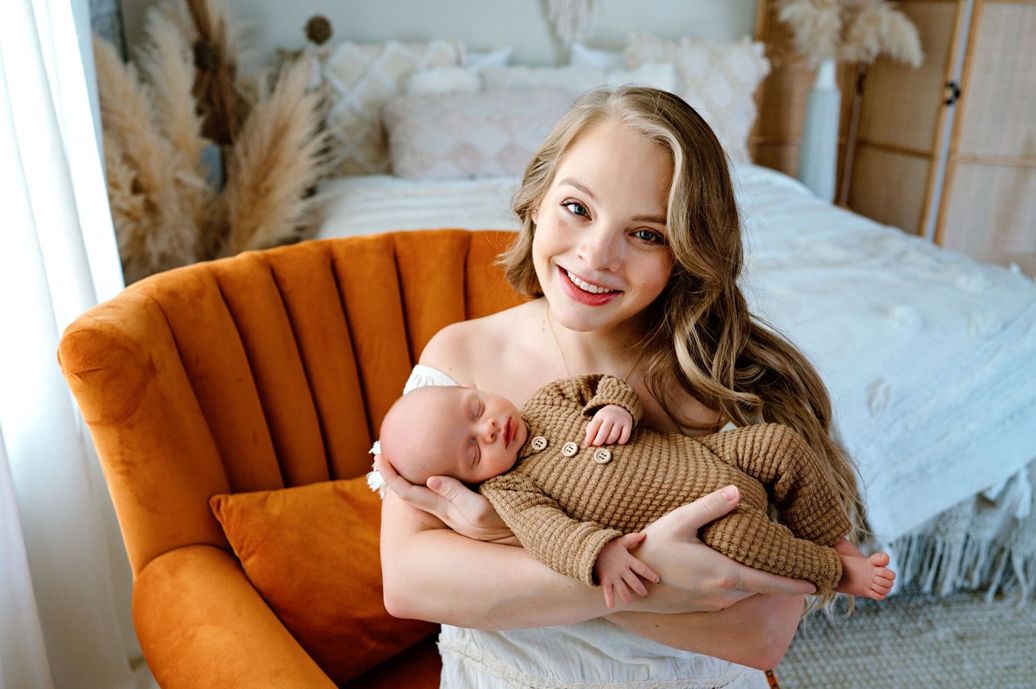 Woman in a beige sweater poses on an orange velvet armchair in a bright white bedroom setting.