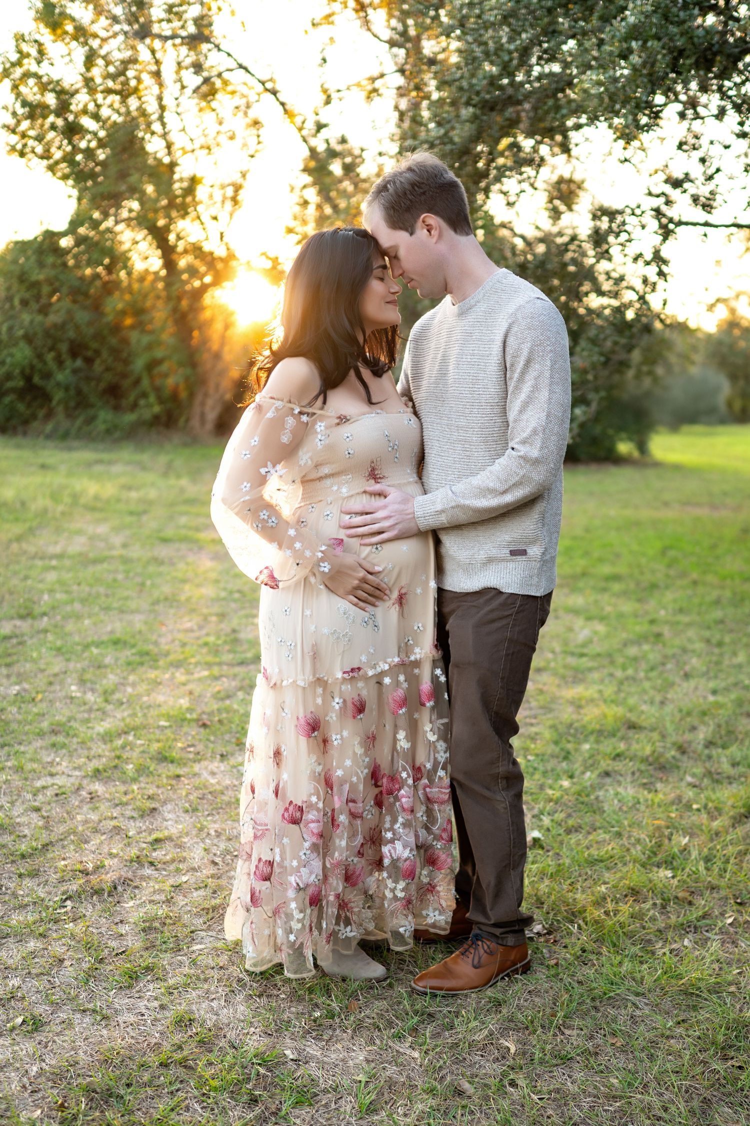 A couple shares an intimate moment at sunset in a field, with the woman wearing a floral dress and man in a sweater.