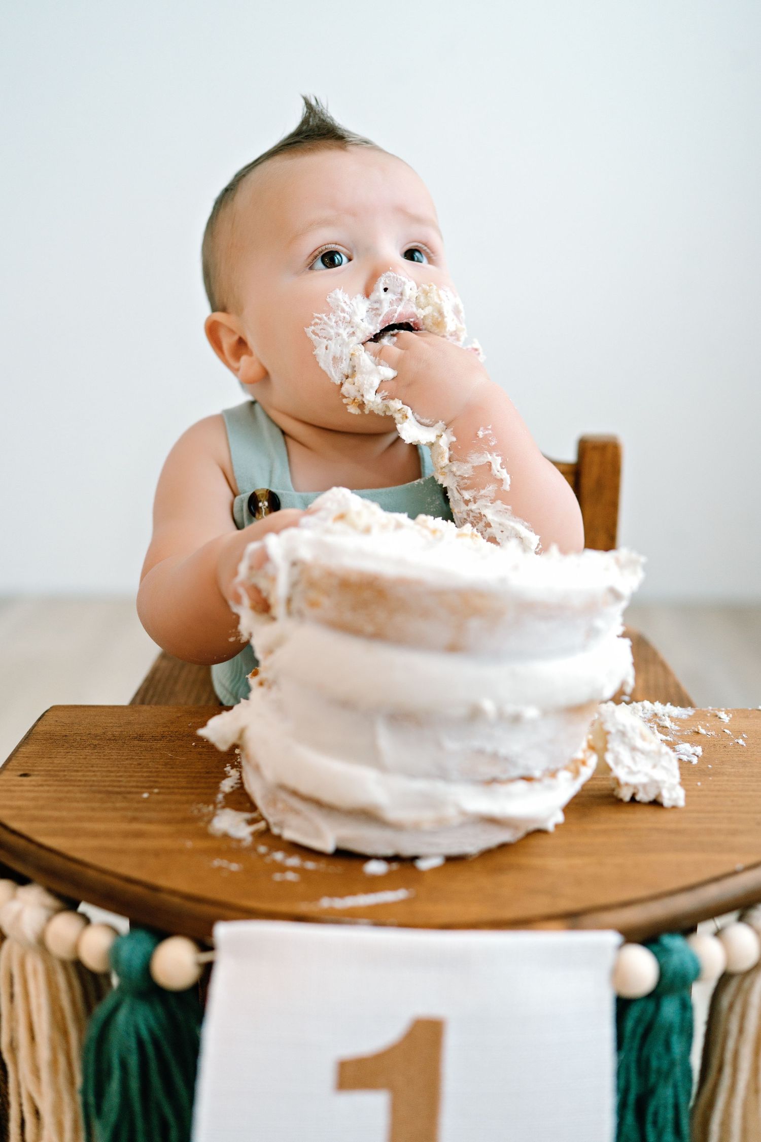 Baby in green outfit dives face-first into white birthday cake on wooden high chair in progression sequence.