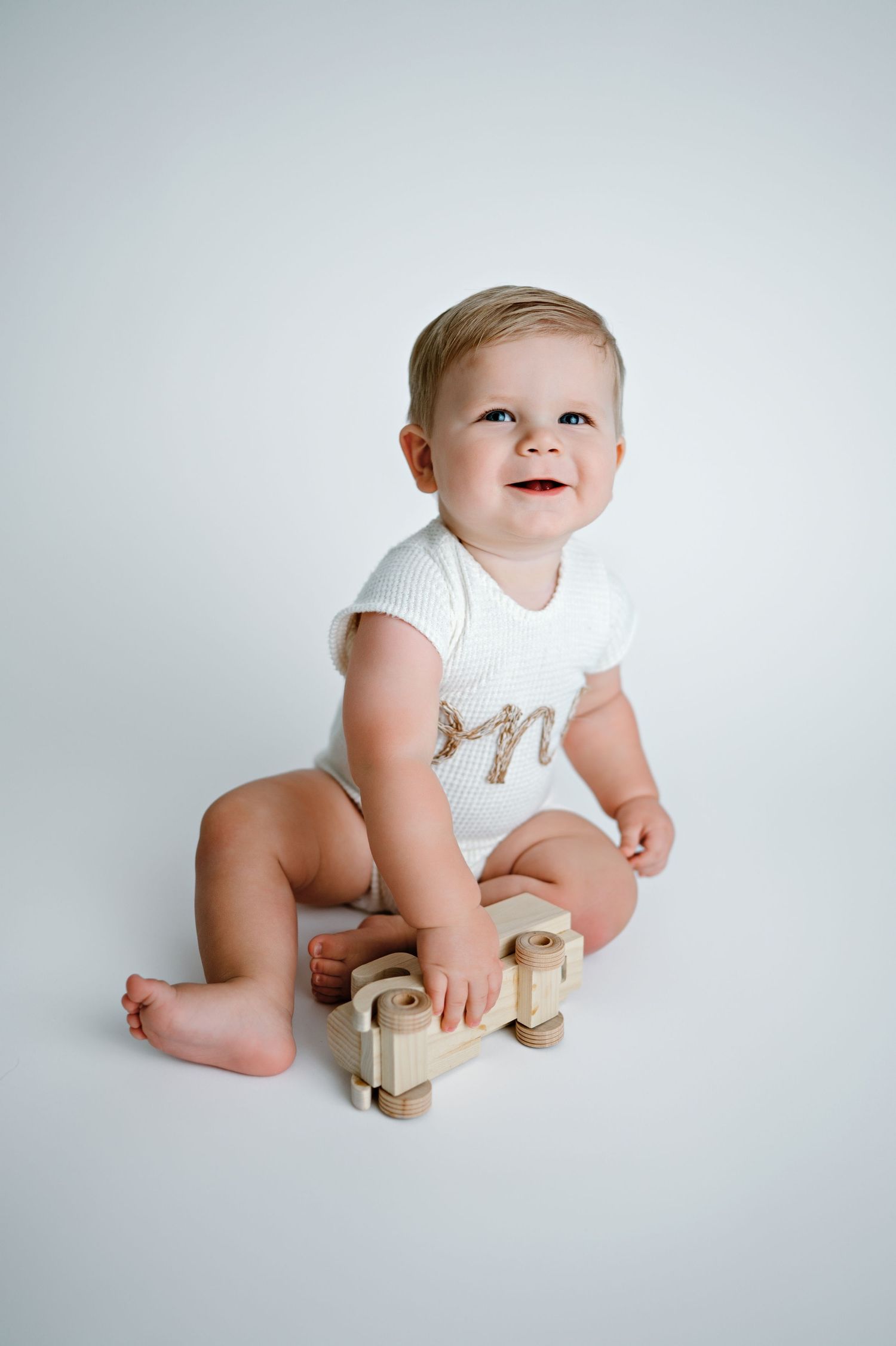 Baby in white onesie plays with wooden toys while sitting on floor against light gray background in progression photos.