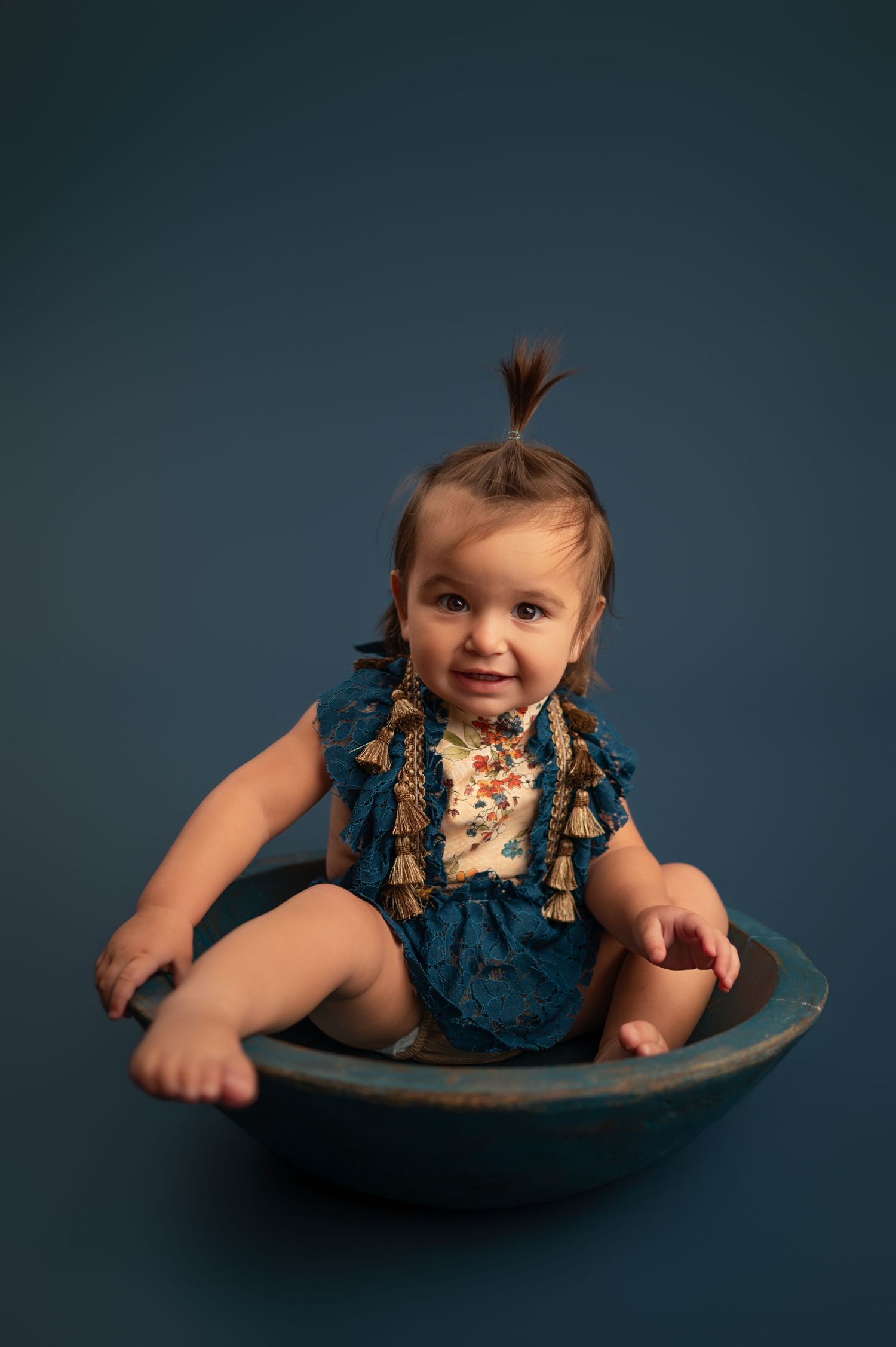 A baby in a navy blue romper sits and plays in a rustic metal bowl against a dark background.