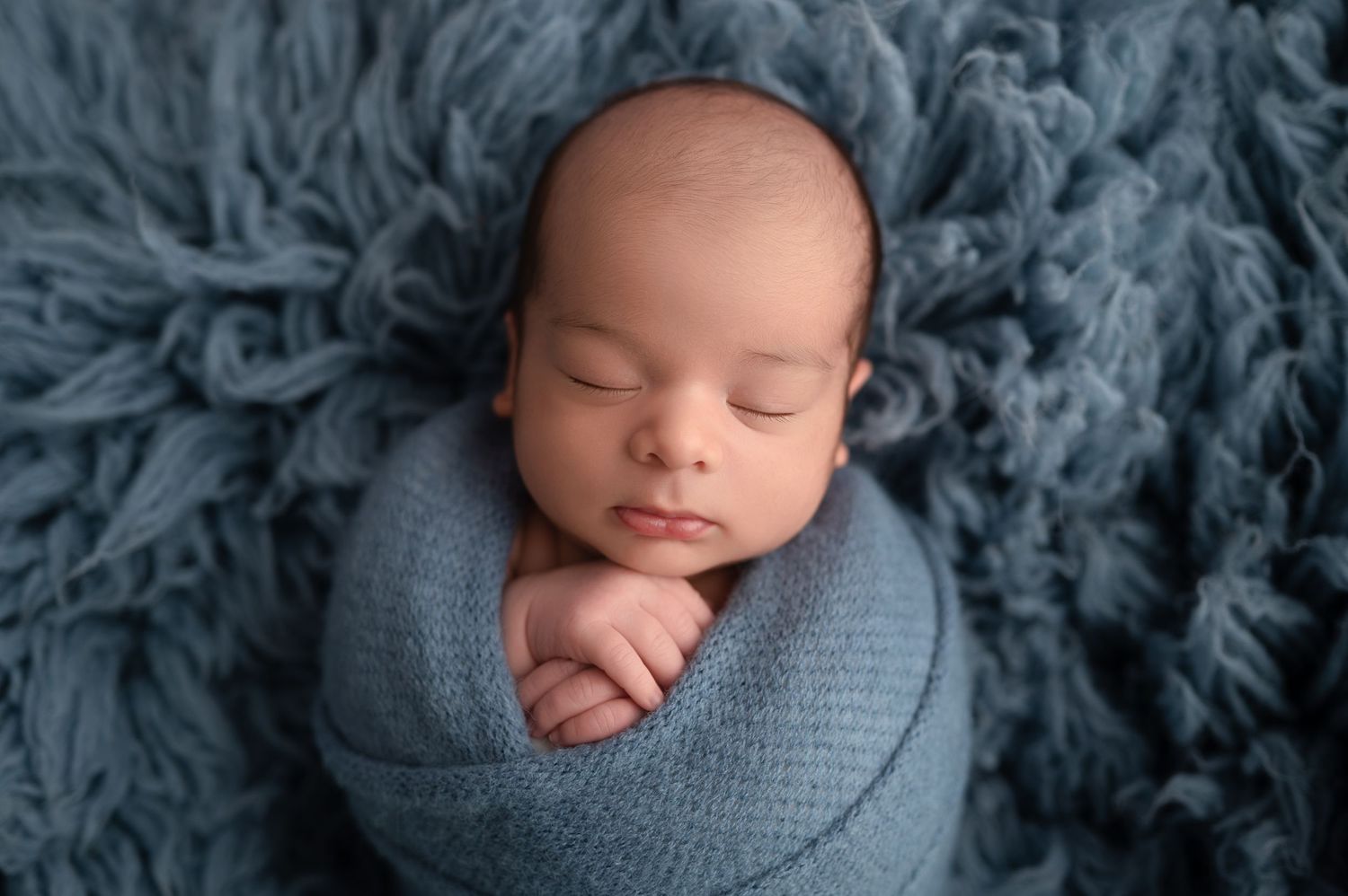 Newborn baby swaddled in blue wrap sleeping peacefully on a soft blue flokati rug in a studio photo.