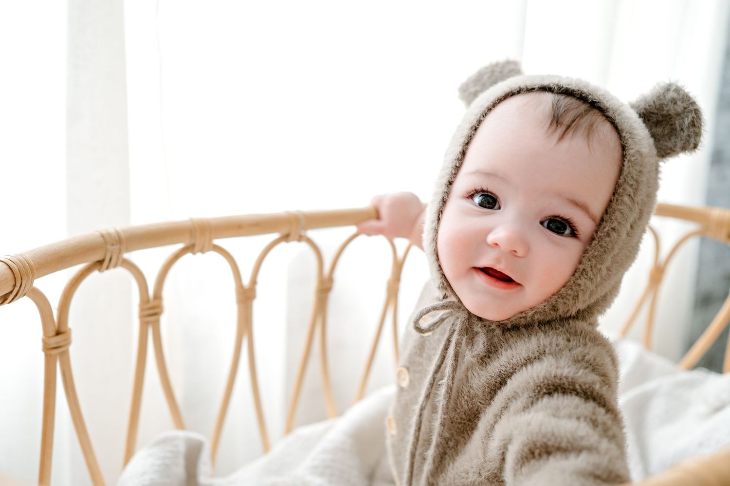 Baby wearing cozy bear hoodie lies on white bedding in natural light.