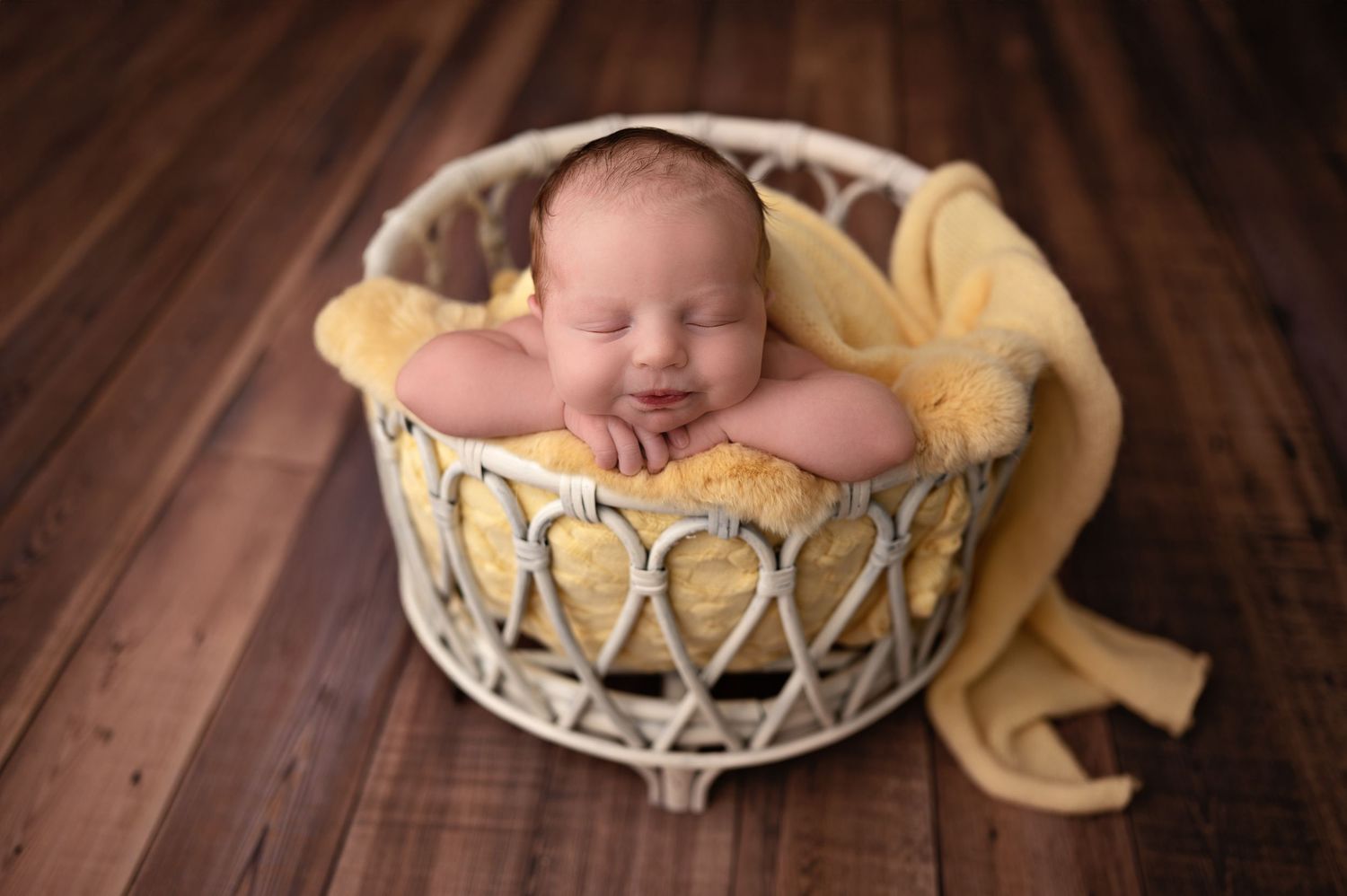 A sleeping newborn baby wrapped in yellow fabric rests peacefully in a white basket on a wooden floor.