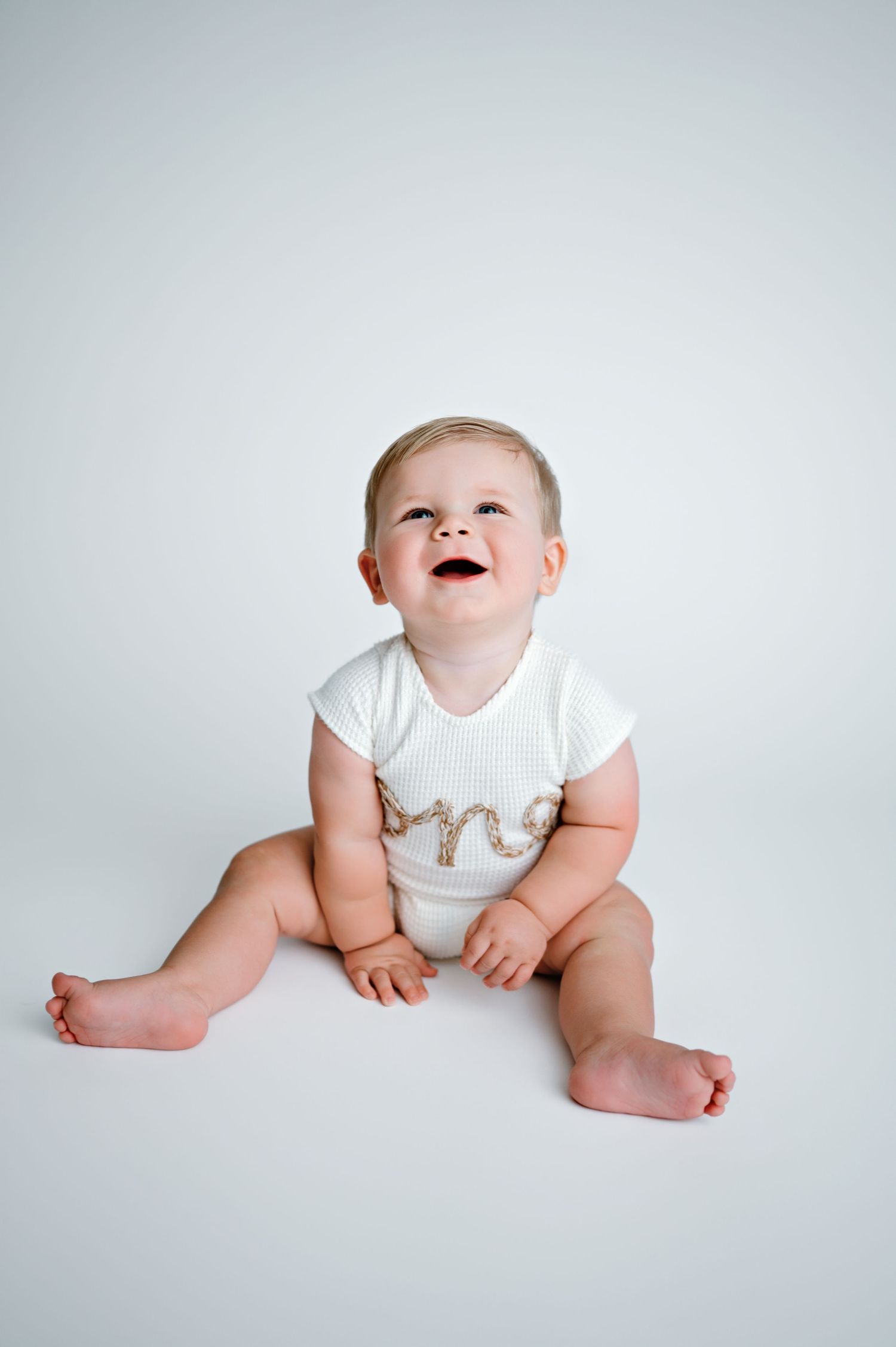 Sweet baby in white onesie sitting and laughing against light gray backdrop in studio photo sequence.