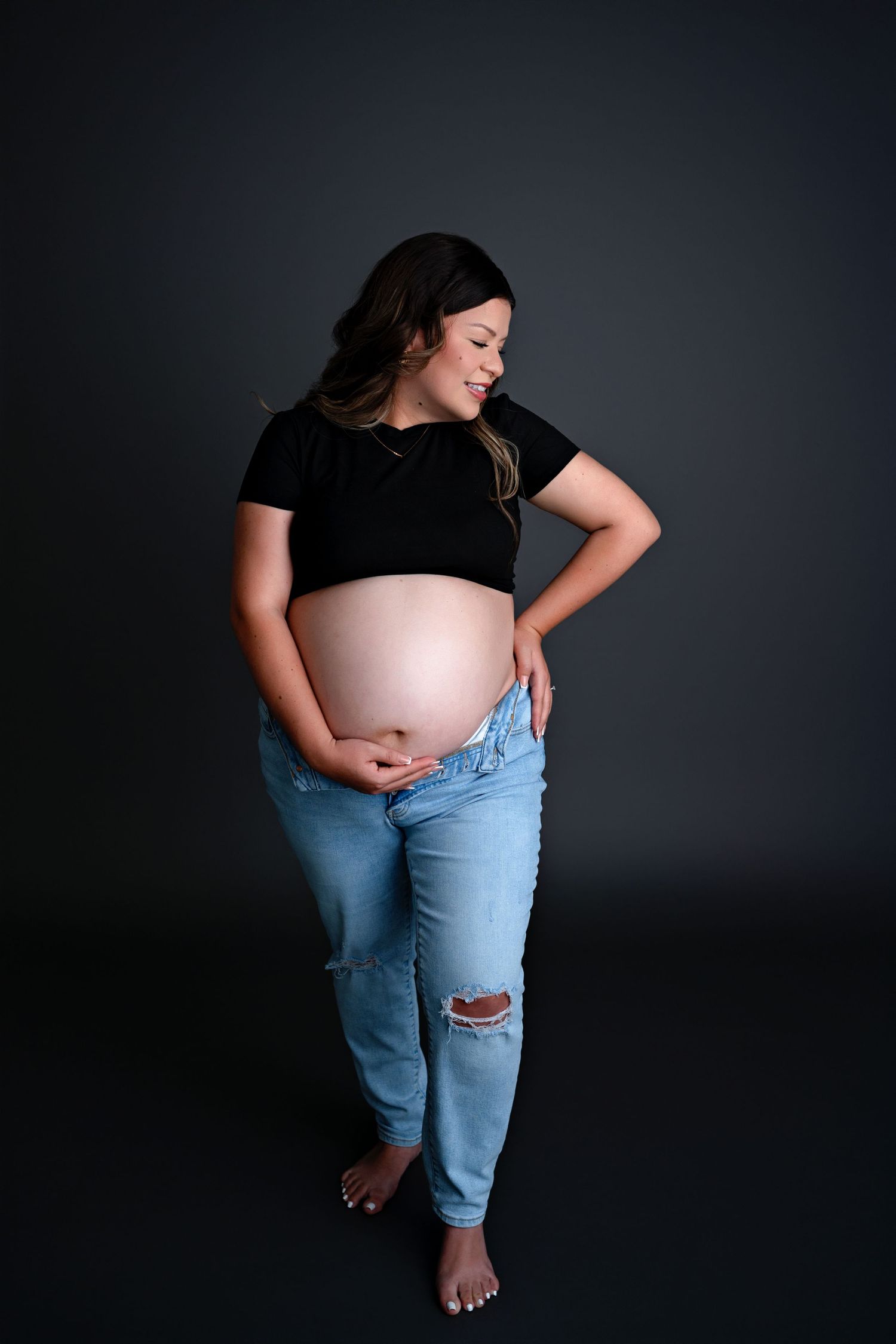 Pregnant woman in black crop top and ripped jeans poses against dark backdrop during maternity photoshoot.