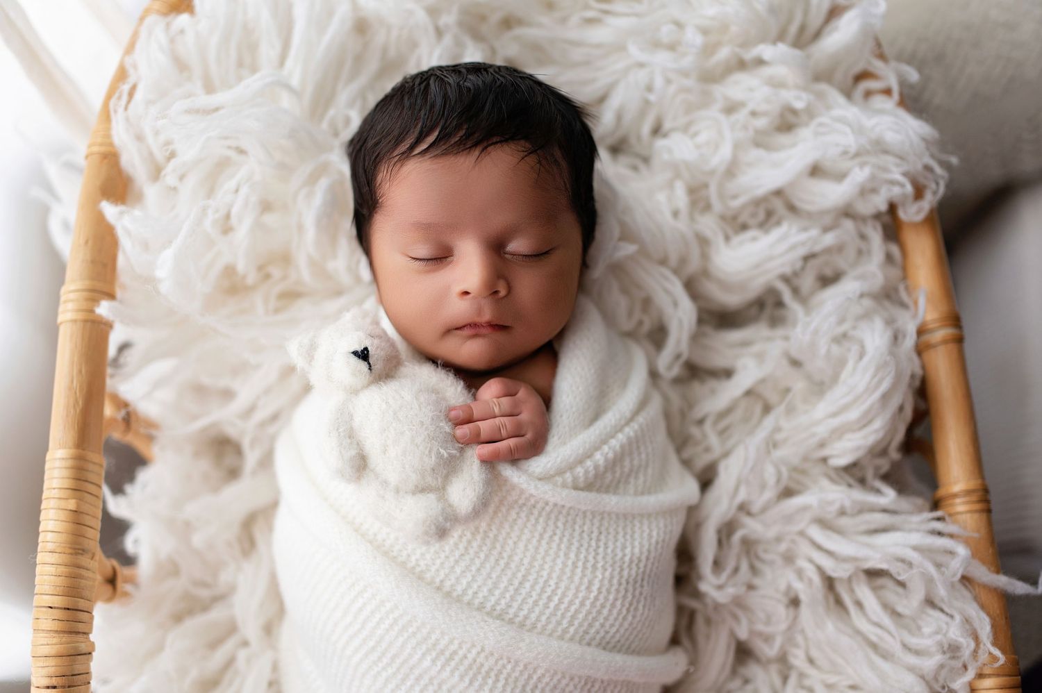 Sweetly sleeping baby swaddled in white on plush fur with wooden bassinet frame visible.