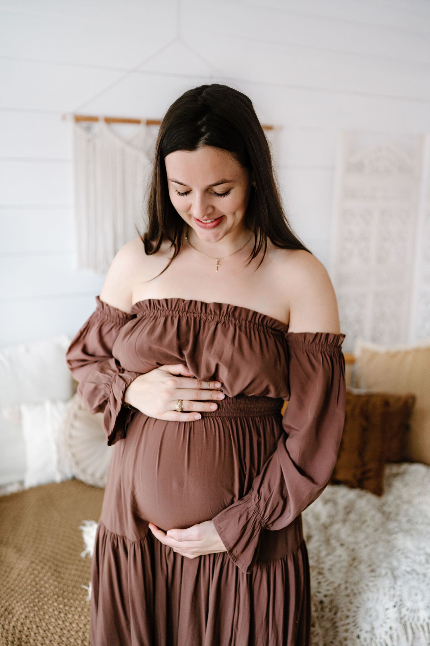 Expectant mother in brown off-shoulder dress with flowing sleeves gently cradles her baby bump in a bright room.
