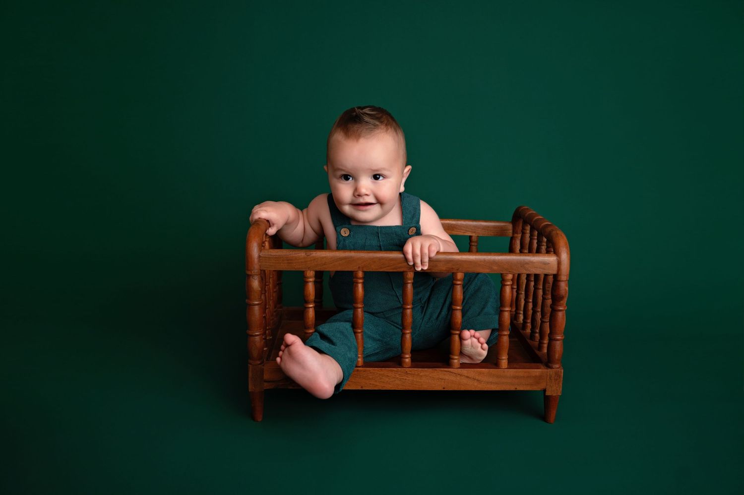 Baby sitting in vintage wooden doll crib against deep green backdrop during professional photo session.