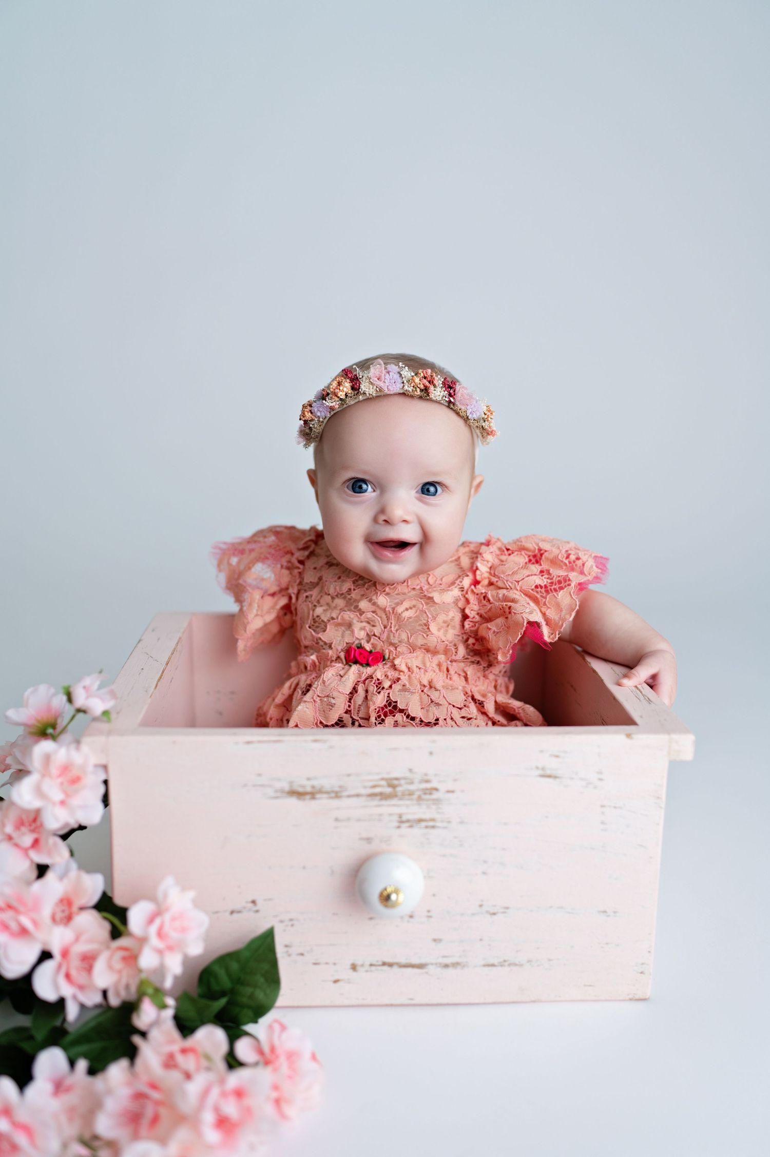 Smiling baby in coral colored outfit poses in vintage white box with pink floral decorations.