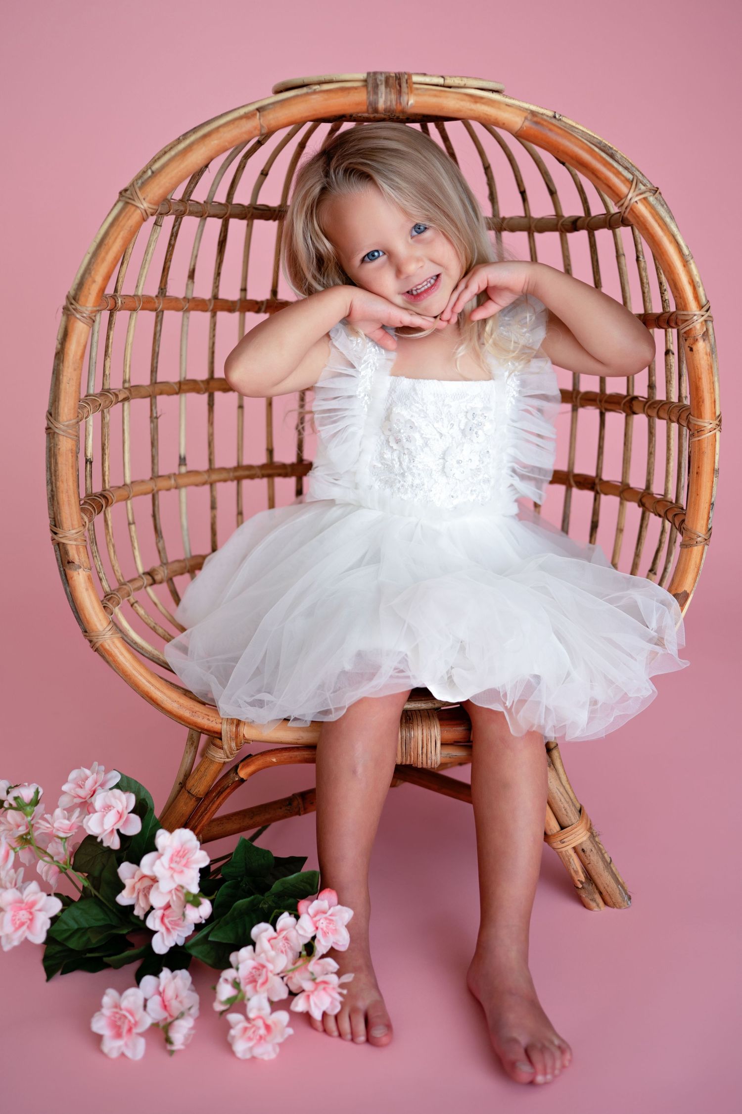 A playful figure wearing a white tulle dress sits in a rattan peacock chair with pink flowers beside it against a pink backdrop.