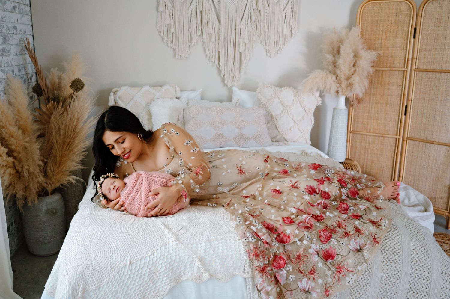 A dreamy photograph showing a woman in a floral dress lounging with a newborn baby on a white bed with boho decor.