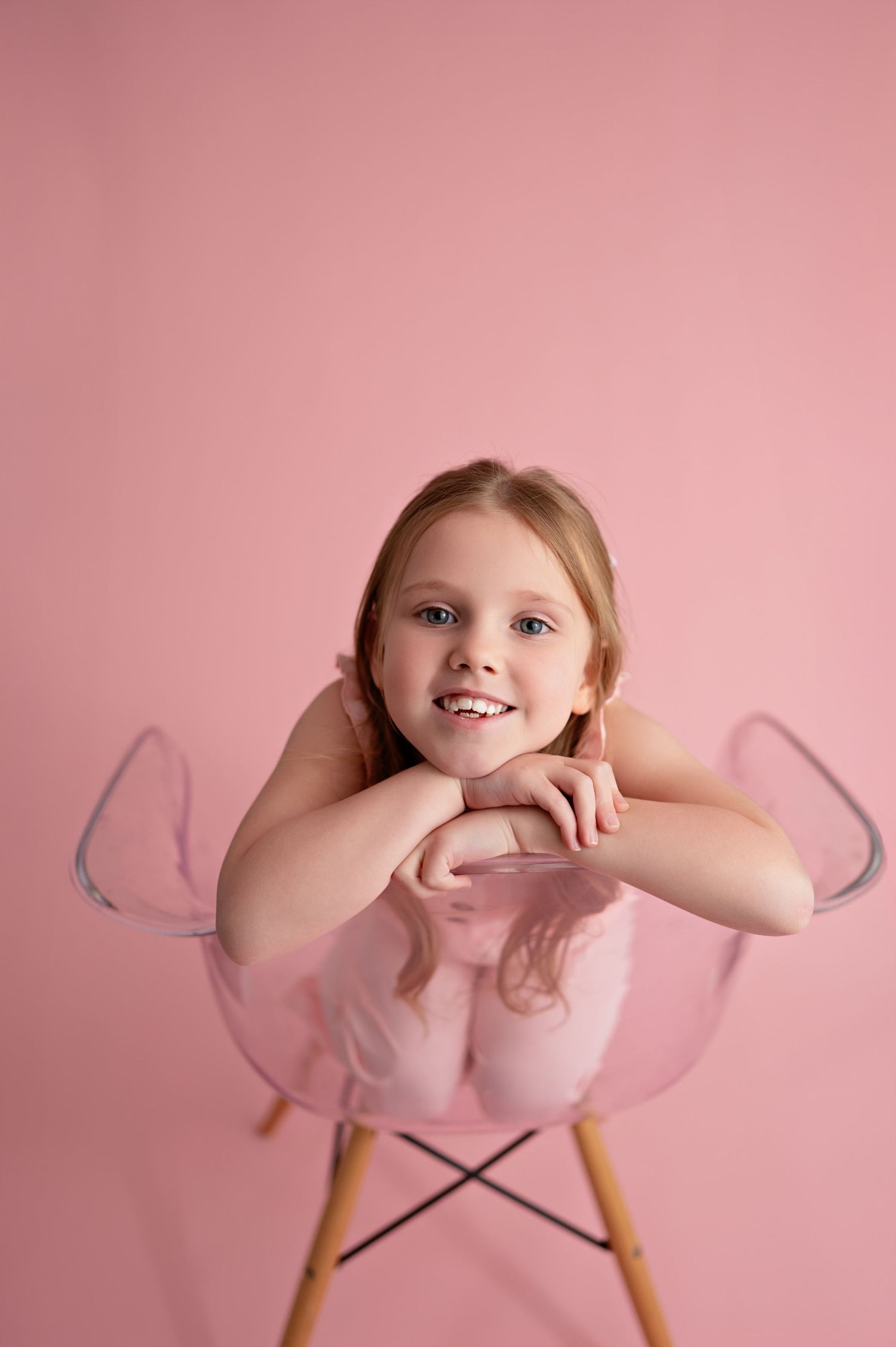 A young child sits in a modern pink Eames-style chair against a pink backdrop in a minimalist portrait setting.