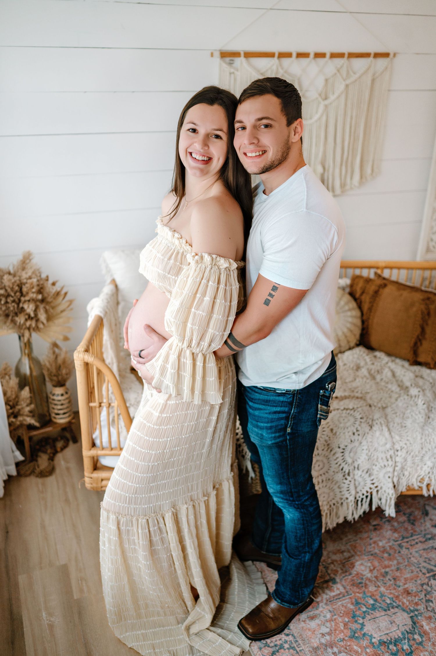A couple poses together in a cozy room with boho decor, white macrame hangings and vintage rugs.