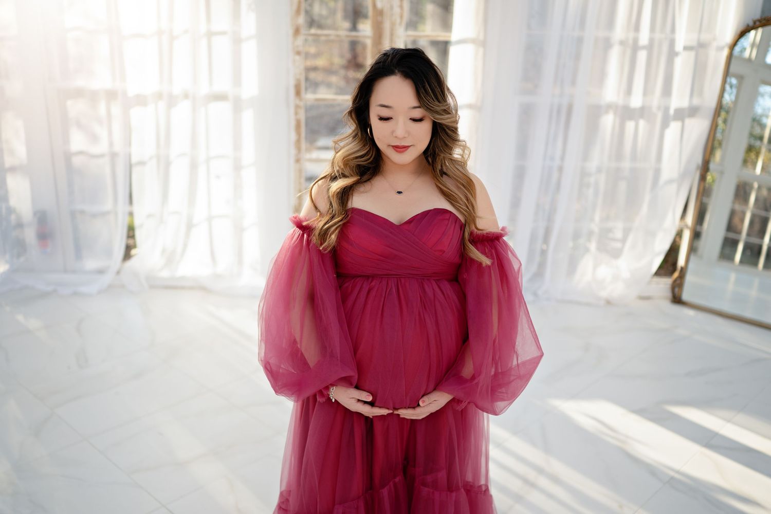 A pregnant woman in a burgundy tulle gown poses in front of sheer white curtains in a sunlit room.