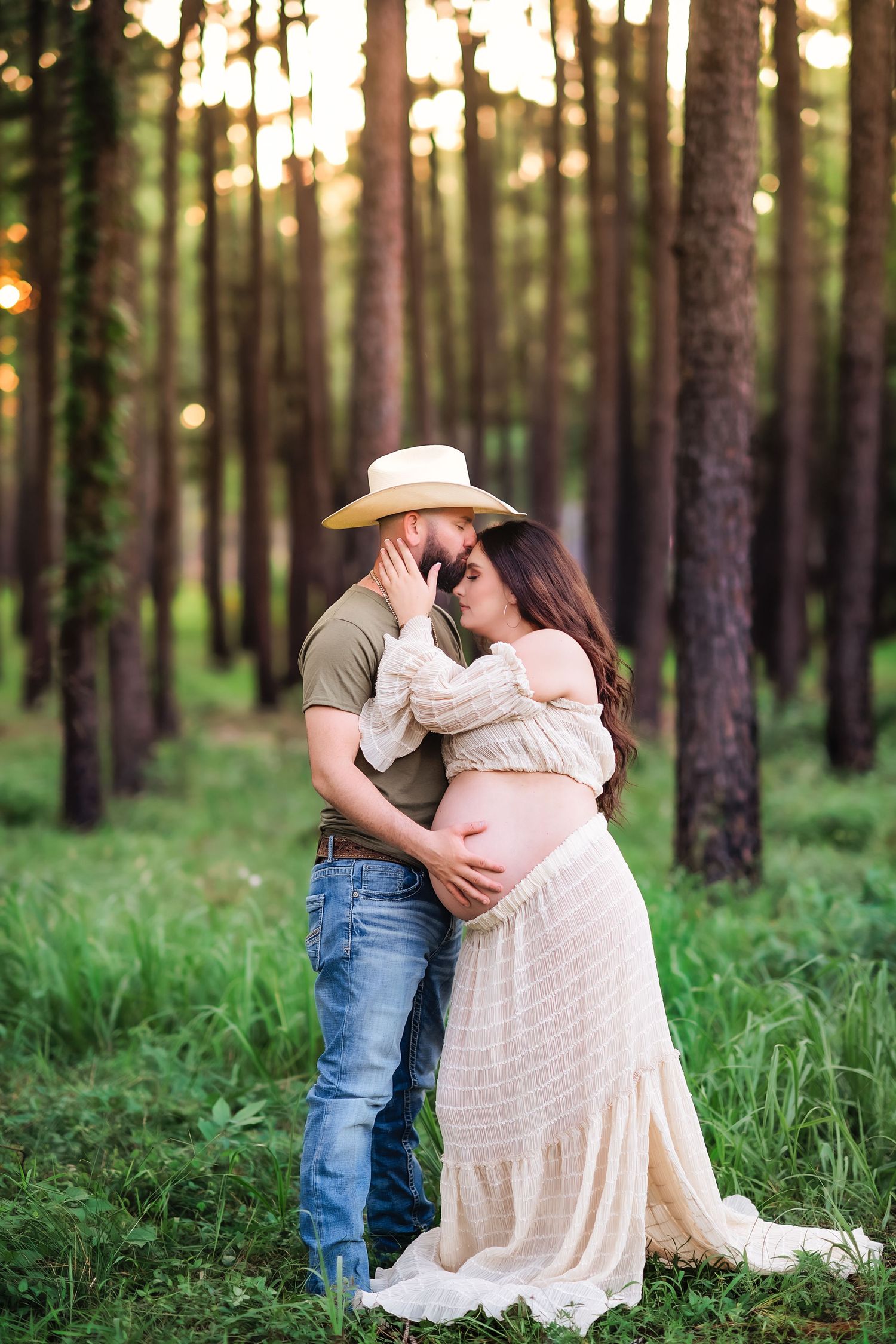 Romantic maternity photo of couple embracing in sunlit forest setting.